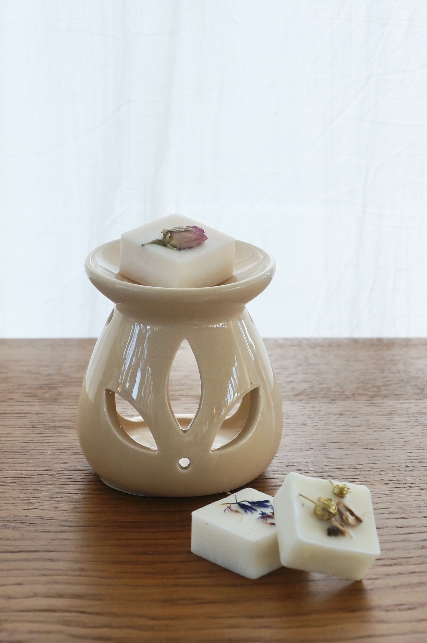 Aromalampa i porslin. Beige. Bild tagen framifrån. En doftpralin ligger i skålen på lampan. Lampan står på ett bord av trä. Vit gardin i bakgrunden.
