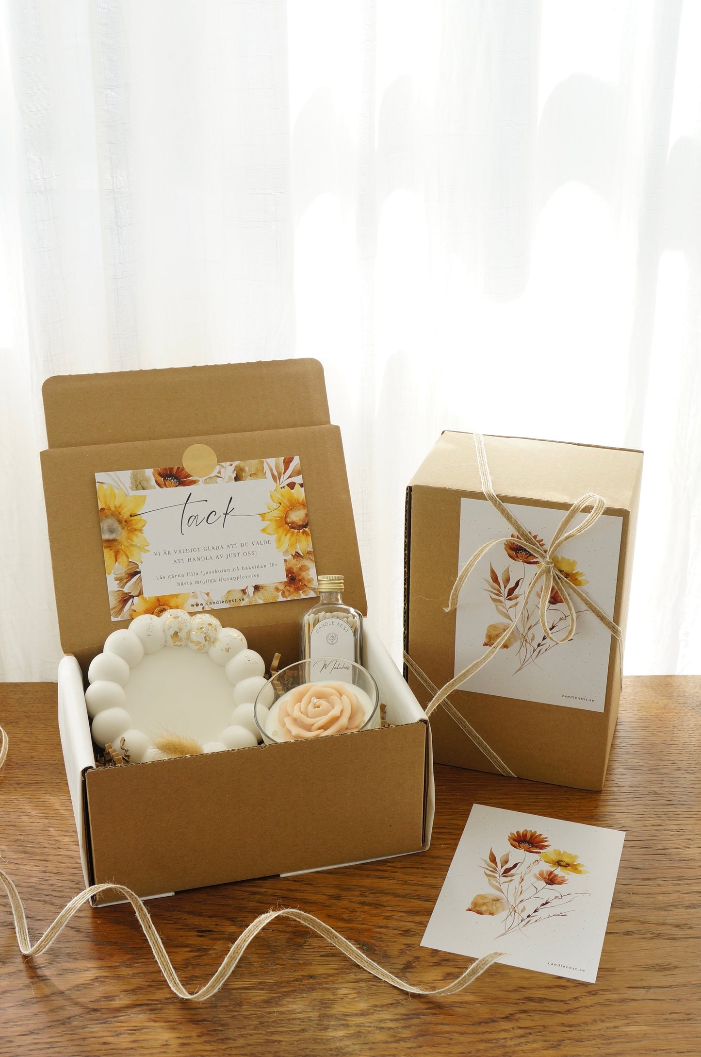 Lyxig Presentbox med ljusfat, Booms & Bubbles doftljus (Pomelo mimosa), flaska med tändstickor. Bild snett ovanifrån med en inslagen presentbox vid sidan