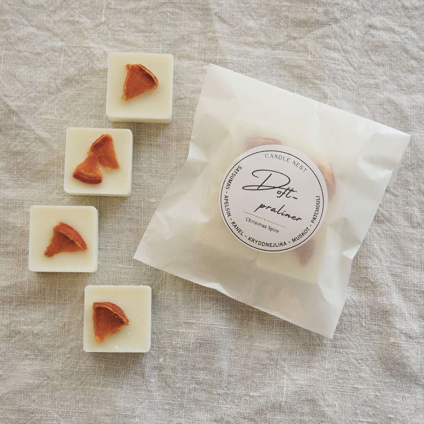En Vit halvtransparent påse innehållandes fyra stycken doftpraliner ligger på en vit duk av linnetyg. Runt påsen ligger fyra stycken doft praliner dekorerade med en gran Grankvist