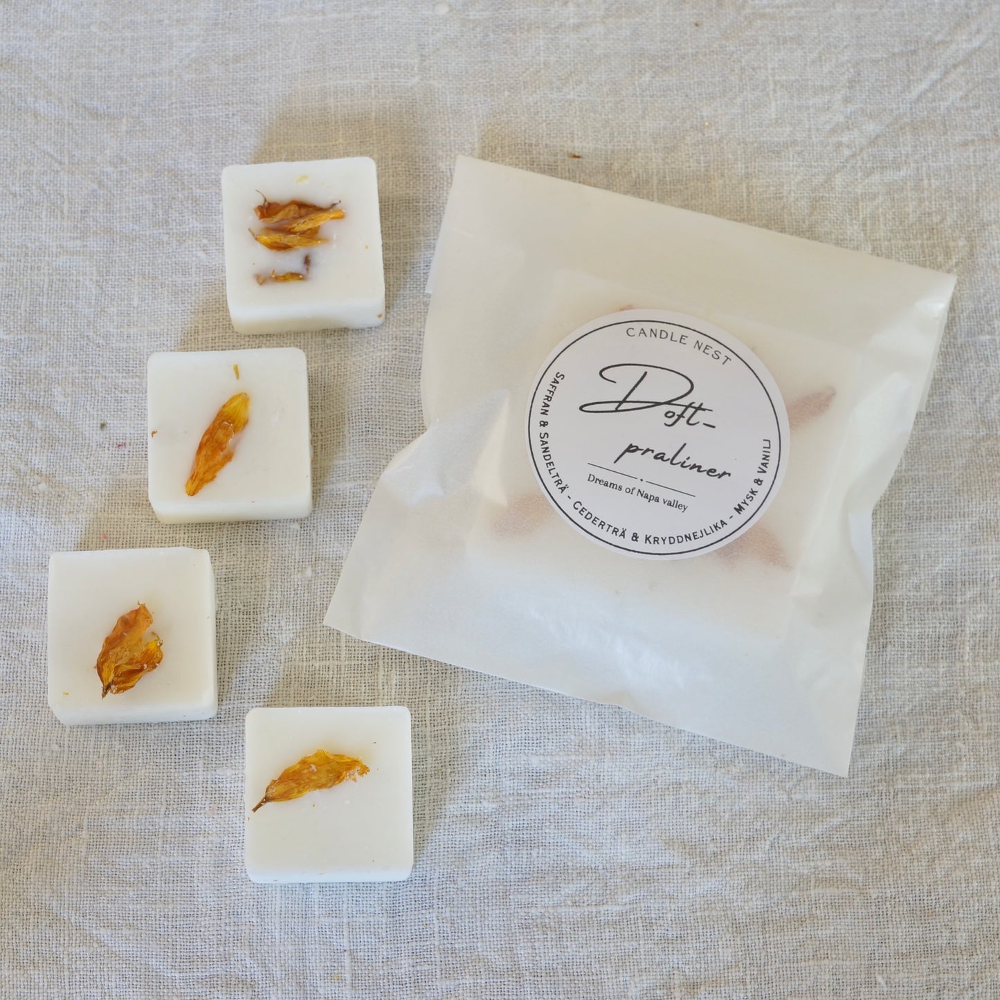 Fyra vita doftpraliner med doften "Dreams of Napa Valley" dekorerade med Torkade kronblad av solros från vår trädgård. Doftvaxen är arrangerade på ett vitt linnetyg bredvid en transparent förpackningspåse från Candle Nest