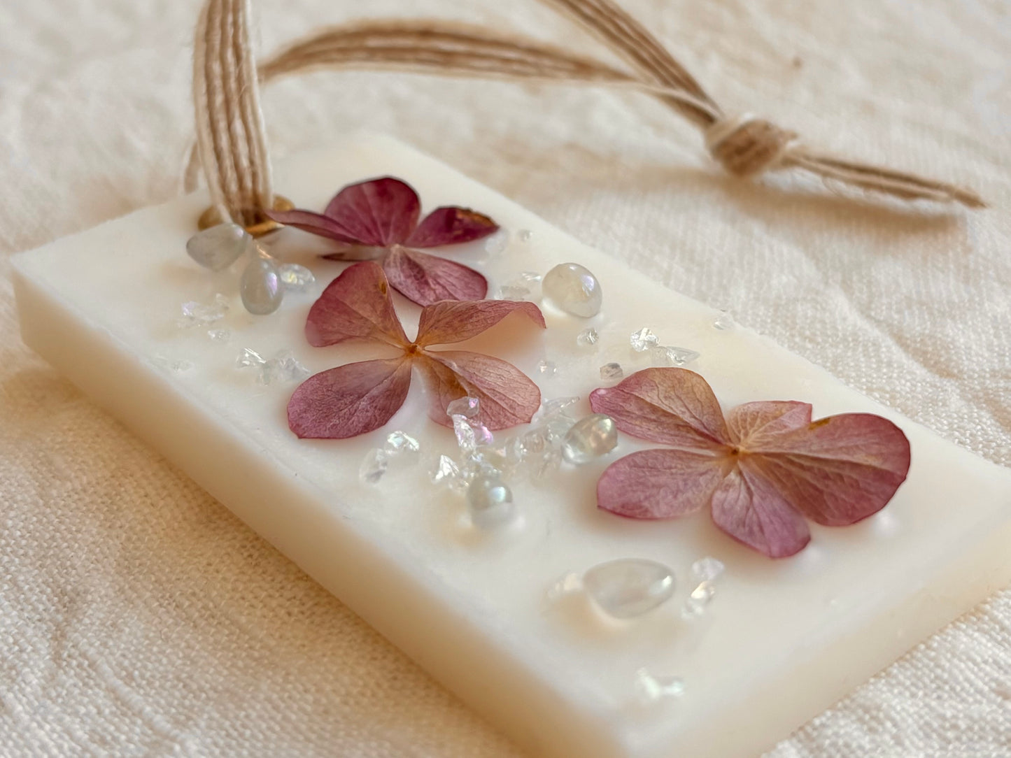 Närbild på Rektangulärt Dofthänge av vax ligger på en vit linneduk. Dofthängen är dekorerat med torkad hortensia och glaspärlor