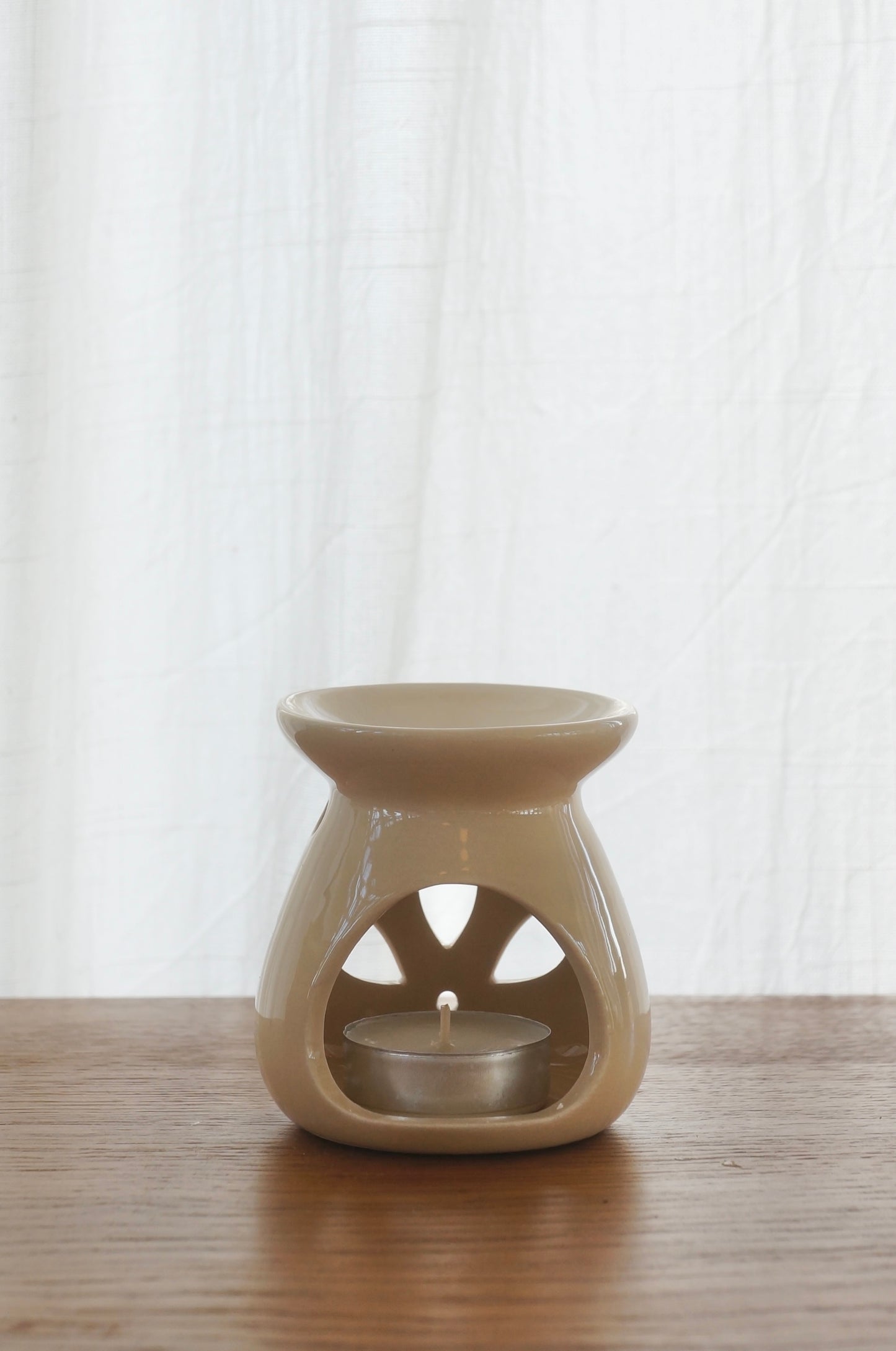 Aromalampa i porslin. Beige. Bild tagen bakifrån. Lampan står på ett bord av trä. Vit gardin i bakgrunden.