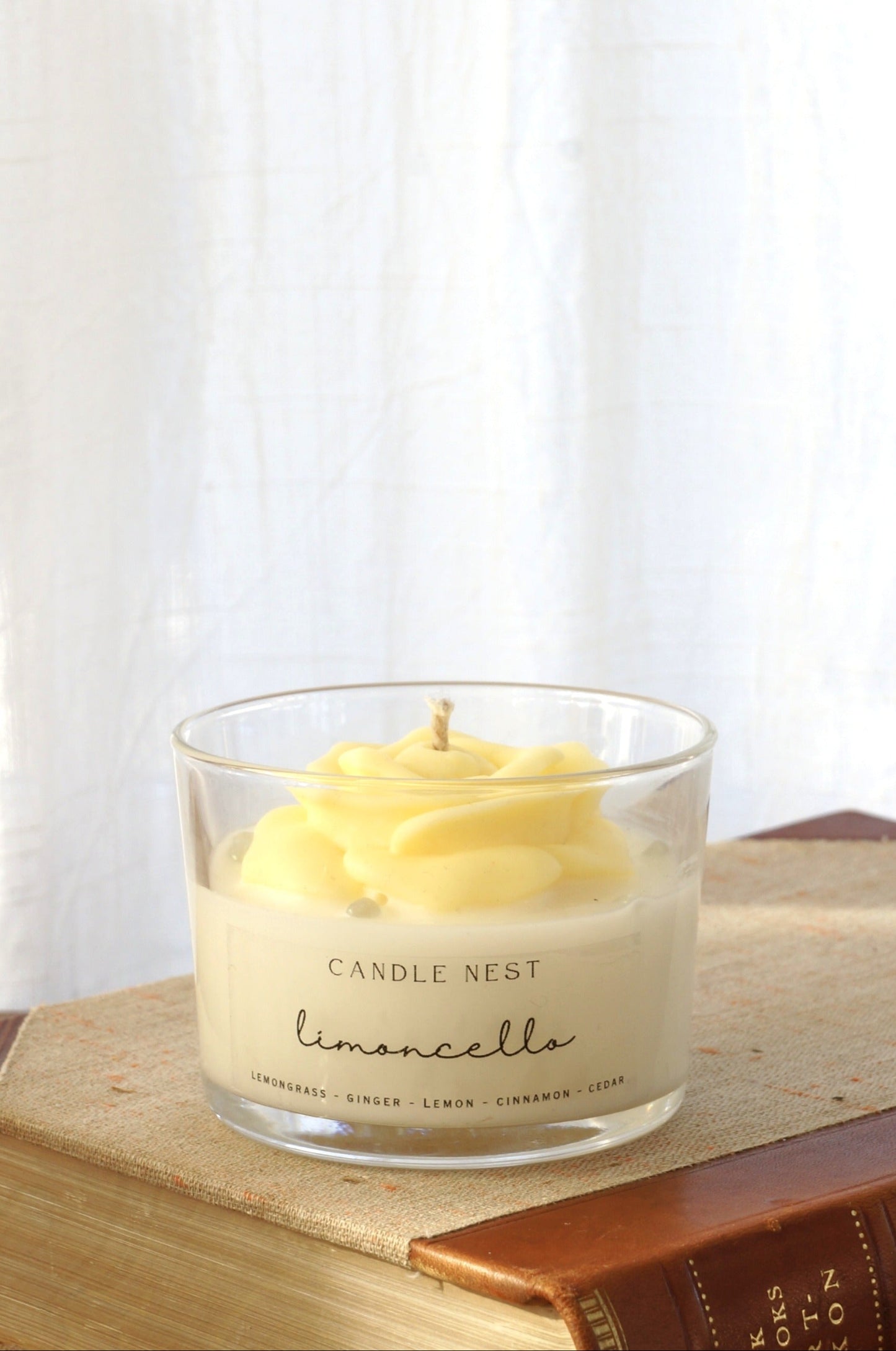 Candle nest doftljus, Limoncello. Transparent glas, en botten av vitt vax och en ljusgul ros av vax på toppen. veke av bomull i mitten av rosen. Ljuset står på en äldre bok. Bild tagen framifrån.