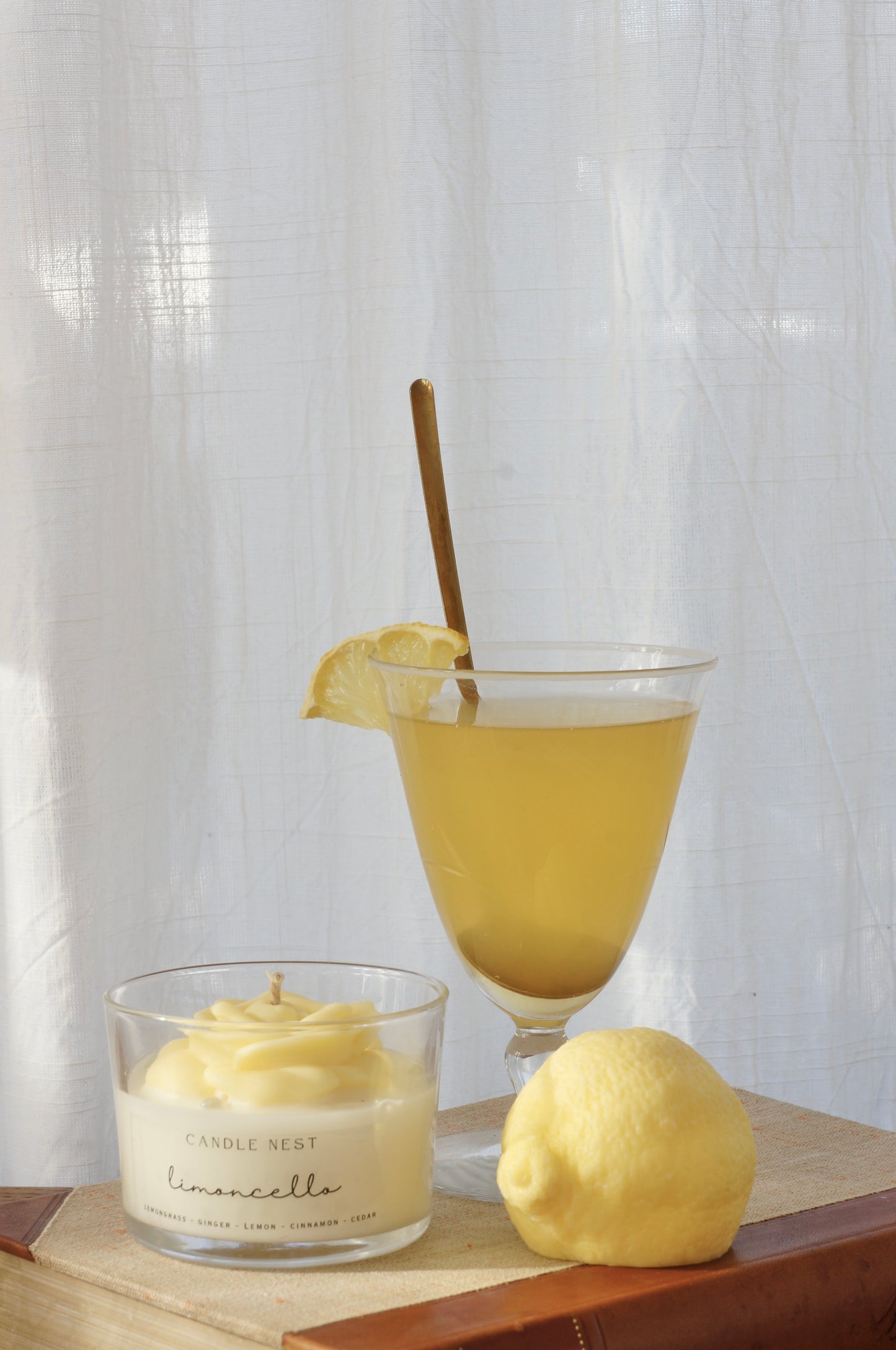 Candle nest doftljus, Limoncello. Transparent glas, en botten av vitt vax och en ljusgul ros på toppen. veke av bomull i mitten av rosen. Ljuset står på en äldre bok och bredvid står en gul lätt transparent drink i ett V-format glas. En en citron ligger bredvid glaset.