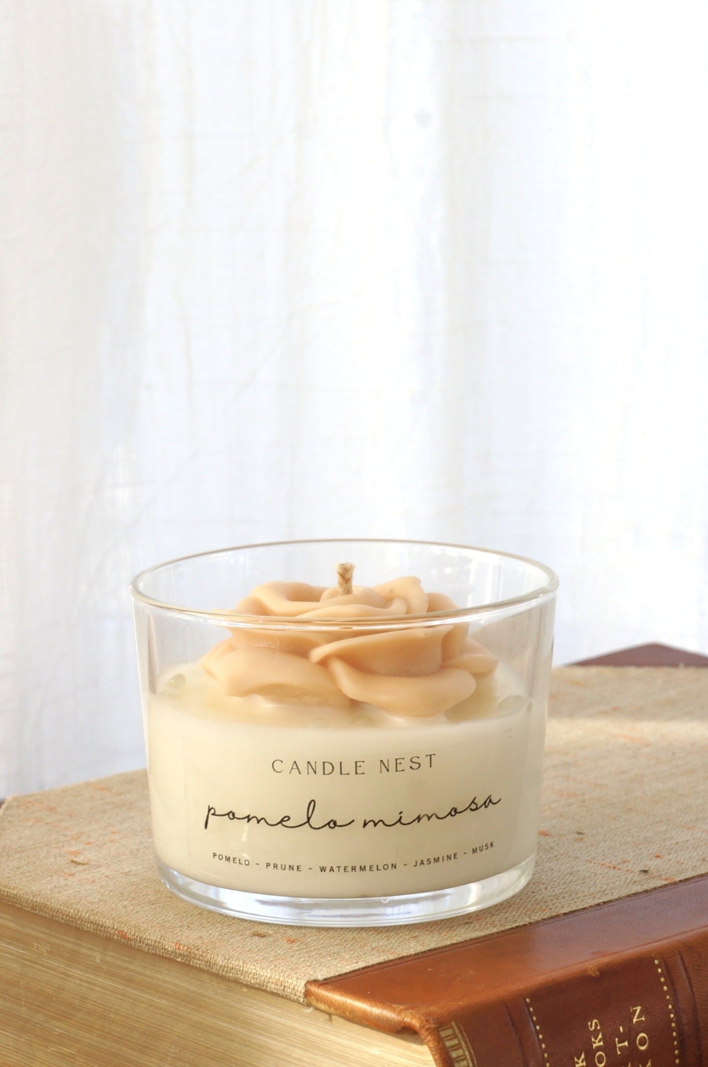 Candle nest doftljus, Pomelo mimosa. Transparent glas, en botten av vitt vax och en ljusrosa ros av vax på toppen. veke av bomull i mitten av rosen. Ljuset står på en äldre bok. Bild tagen framifrån.