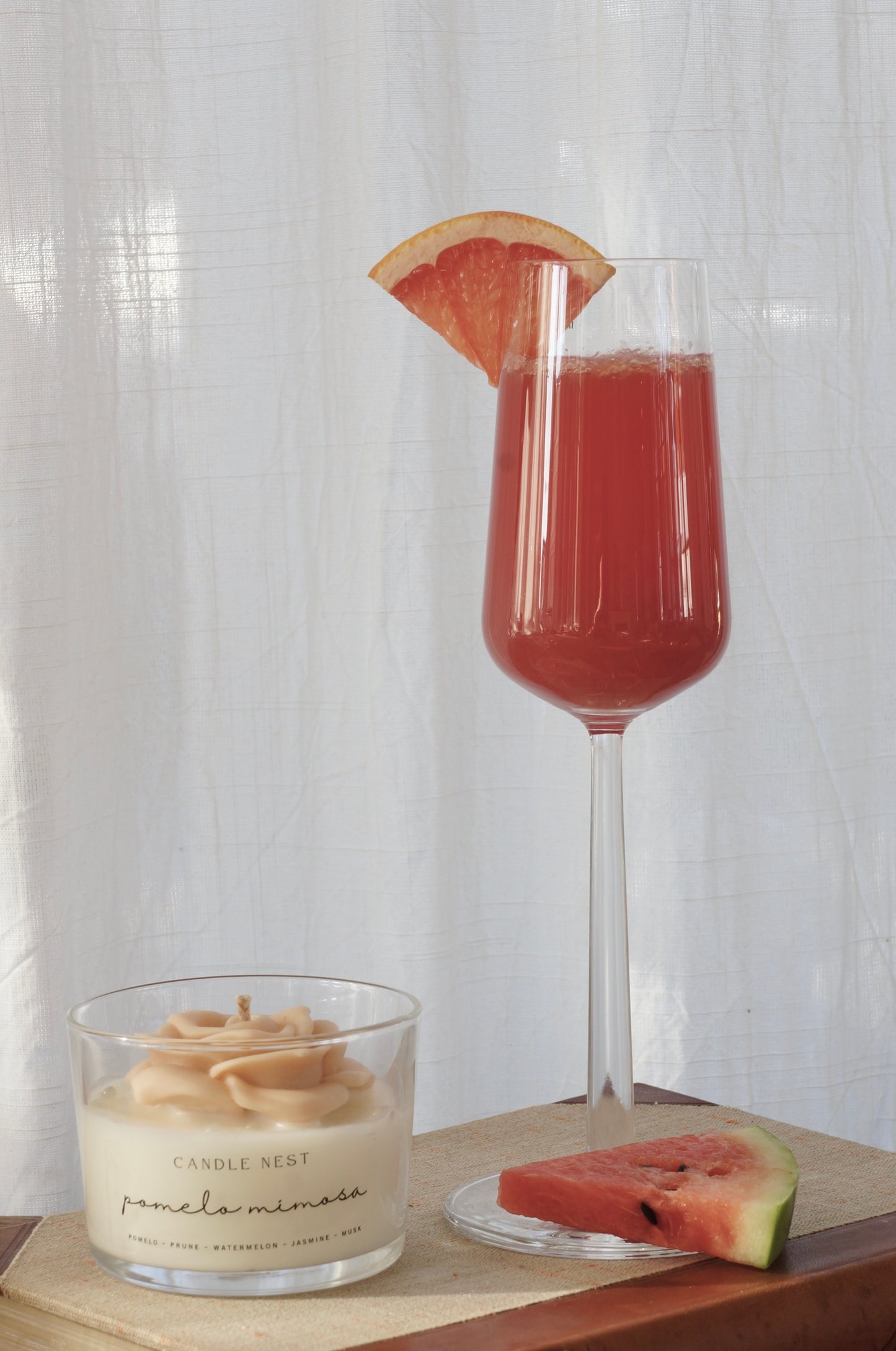 Candle nest doftljus, Pomelo mimosa. Transparent glas, en botten av vitt vax och en ljusrosa ros av vax på toppen. veke av bomull i mitten av rosen. Ljuset står på en äldre bok och bredvid står en röd drink i ett högt glas. En skiva vattenmelon ligger på glasets fot. Bild tagen framifrån.