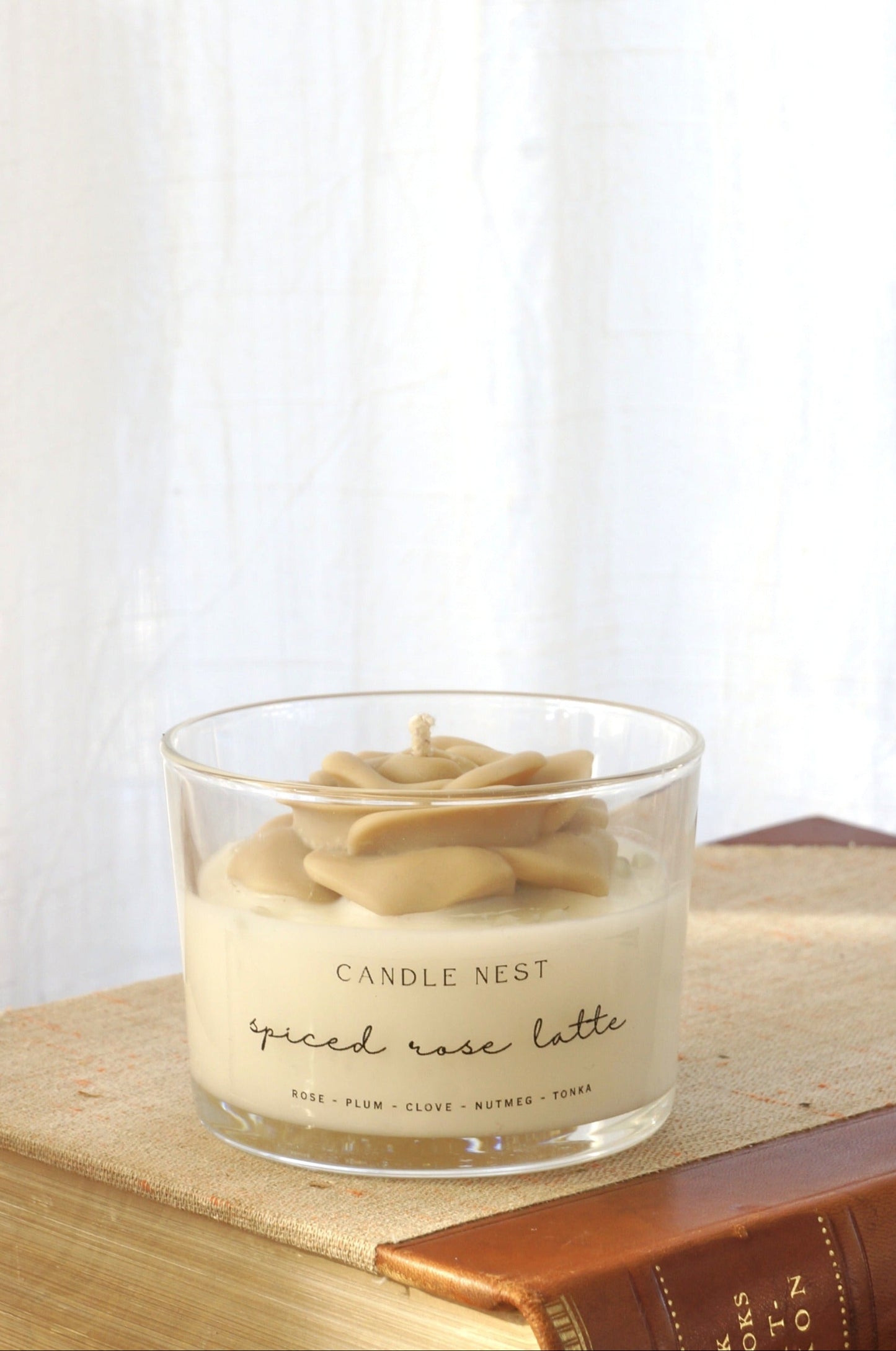 Candle nest doftljus, Spiced rose latte. Transparent glas, en botten av vitt vax och en beige ros på toppen. veka av bomull i mitten av rosen. Ljuset står på en äldre bok som ligger på ett bord av trä. Bild tagen från sidan
