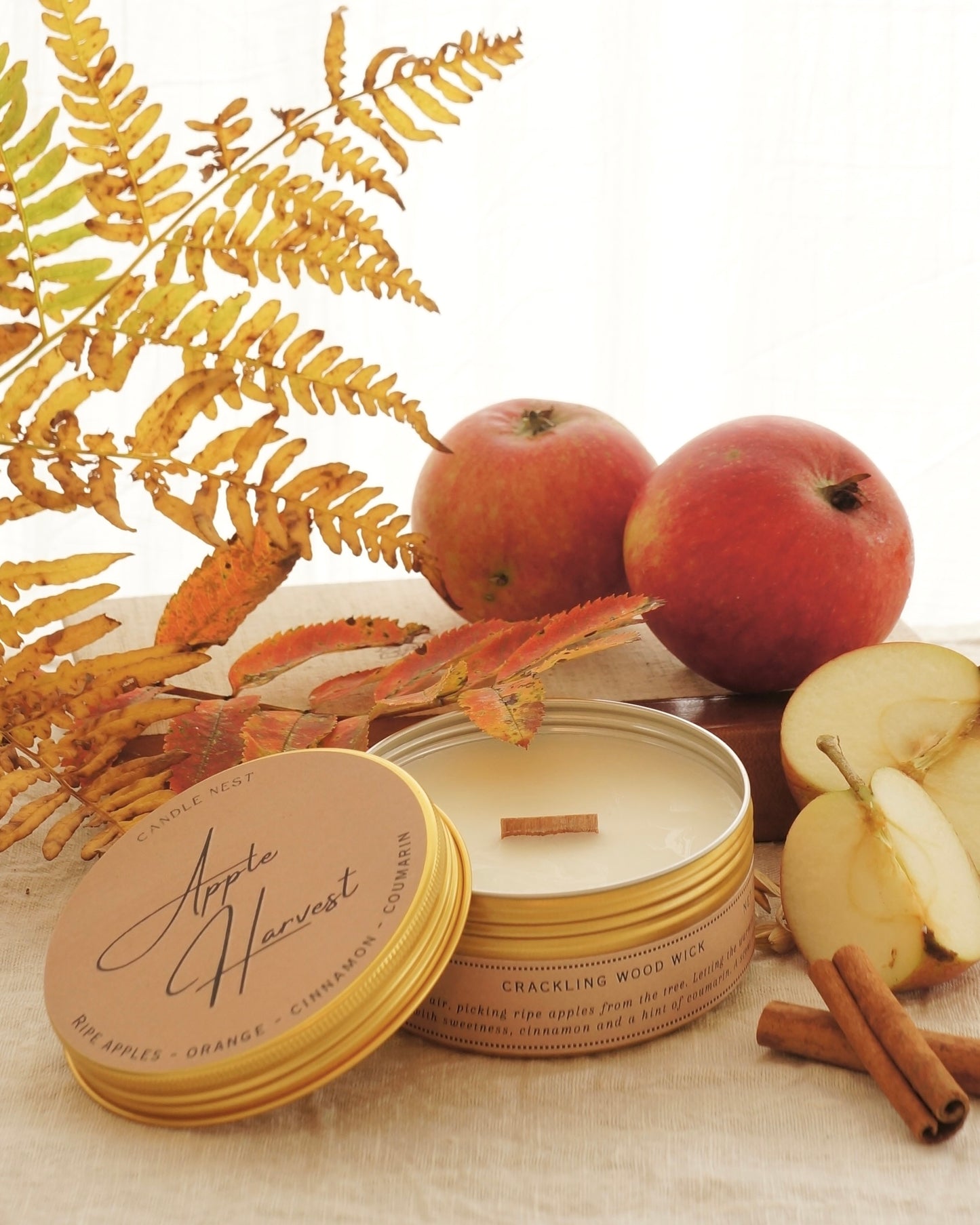 Candle nest doftljus "Apple Harvest" i en rund guldig burk av metall står på ett bord omgivet av äpplen,, kanel och höstliga löv och kvistar. Locket är lutat mot sidan av burken och vax och veke av trä är exponerat.