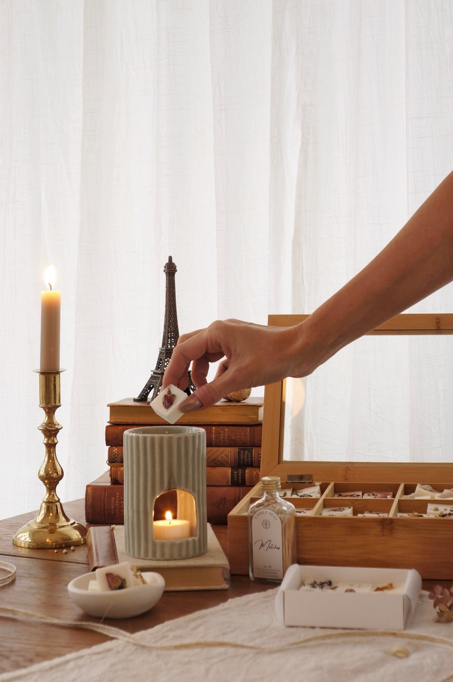 Hand som placerar doftpralin i vit räfflad aromalampa omgiven av tända ljus, vintage-böcker och träbox med doftpraliner i hemtrevlig miljö