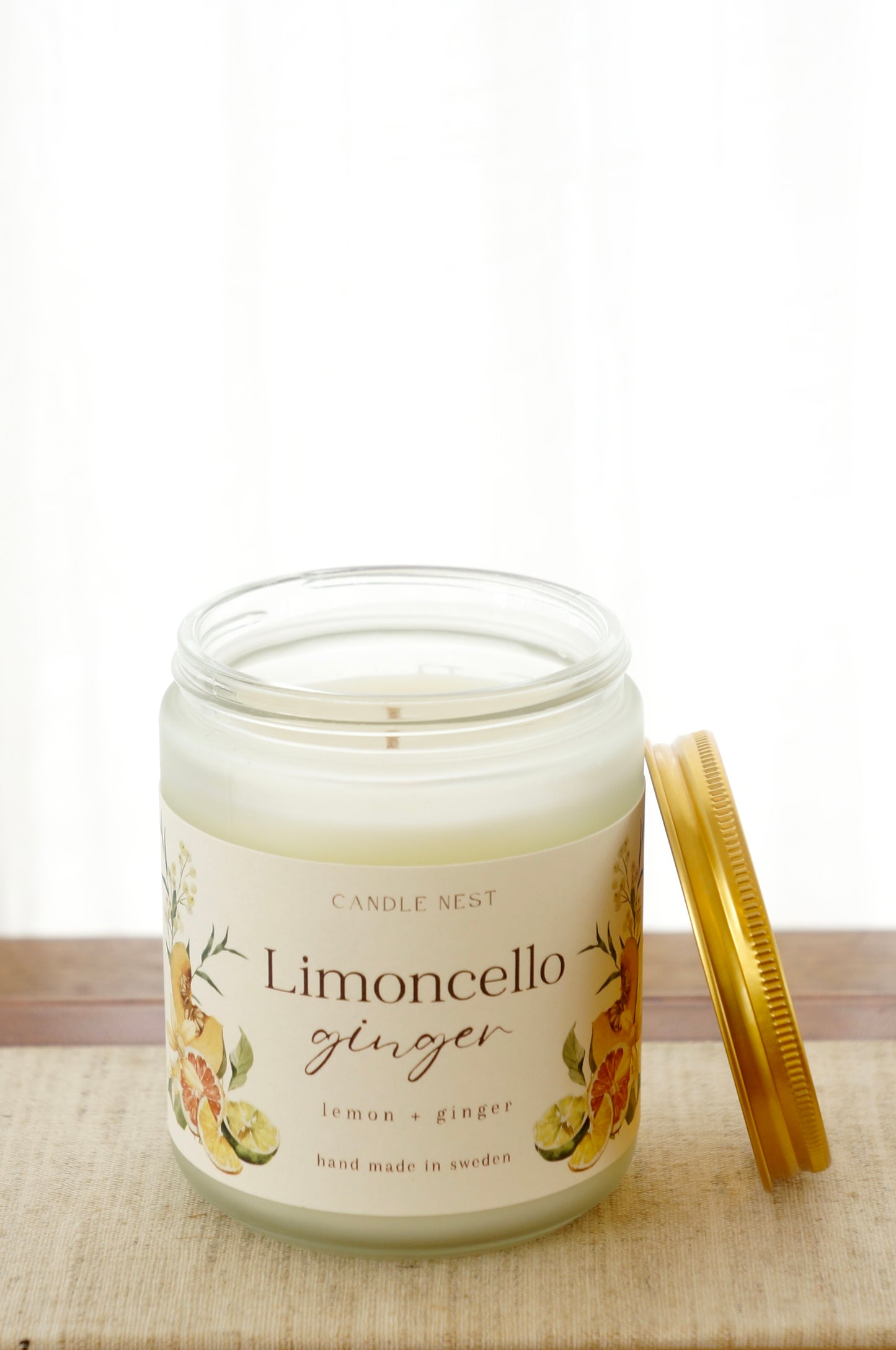 Ett doftljus i en frostad vit glasburk står på en bok. Runt ljuset finns ett off-white papper med bilder av frukter och texten "Limoncello ginger". Mot ljuset står burken skruvlock i guld lutat.