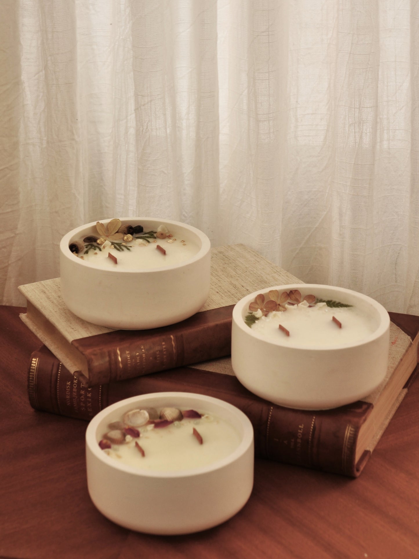 Tre stycken handgjorda doftljus i vita gipsskålar fyllda med rapsvax, sprakande trävekar och mjuka, lyxiga dofter. Dekorerad med torkade blommor. Bild tagen framifrån, snett uppifrån