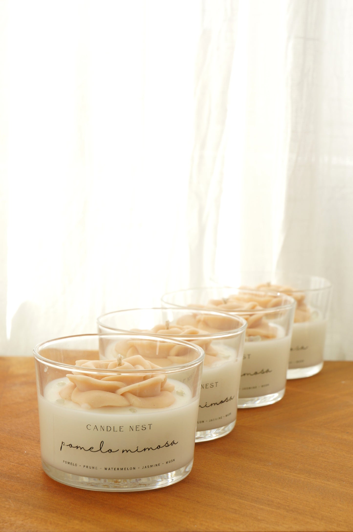 Fyra stycken Candle Nest Pomelo Mimosa glasljus stående i rad med persikofärgad rosdekor som flyter på vitt vax, ståendes på en träyta i naturligt ljus