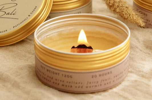 Ett doftljus i en guldig metallburk står på ett bord. Mitt i ljuset syns en bred veke av trä som brinner. Rapsvaxet i burken är nästan helt smält och visar en full smältpool.