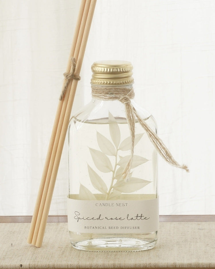 Doftpinnar. Genomskinlig flaska med skruvkork i guld och med en vit bladig växt i. Syntetiska pinnar står lutande mot flaskan. 