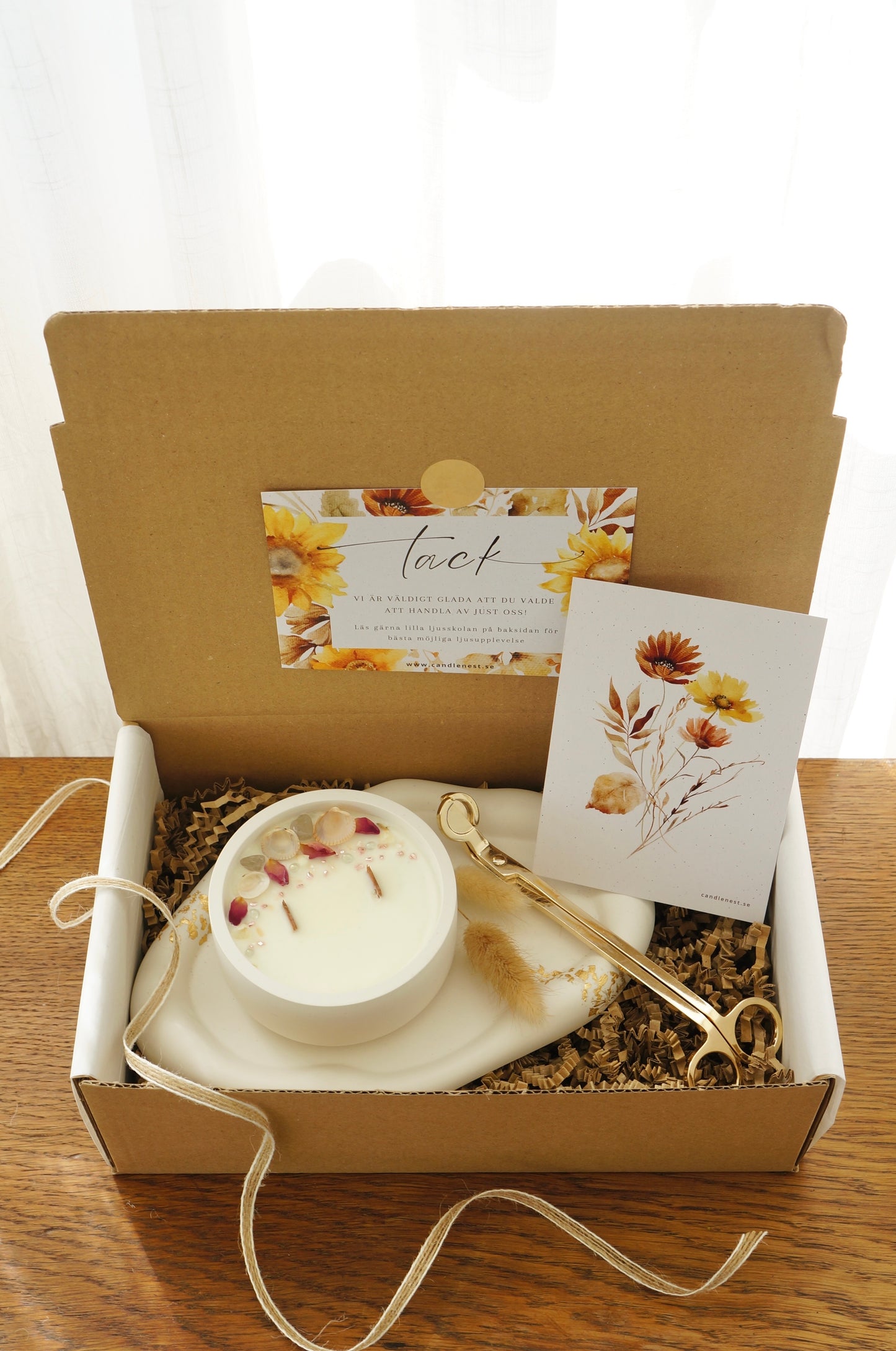 Presentbox med Botaniskt doftljus (sunset at the beach), ljusfat med gyllen dekor och en vektång. Produkterna ligger i en låda av naturfärgd papp. 