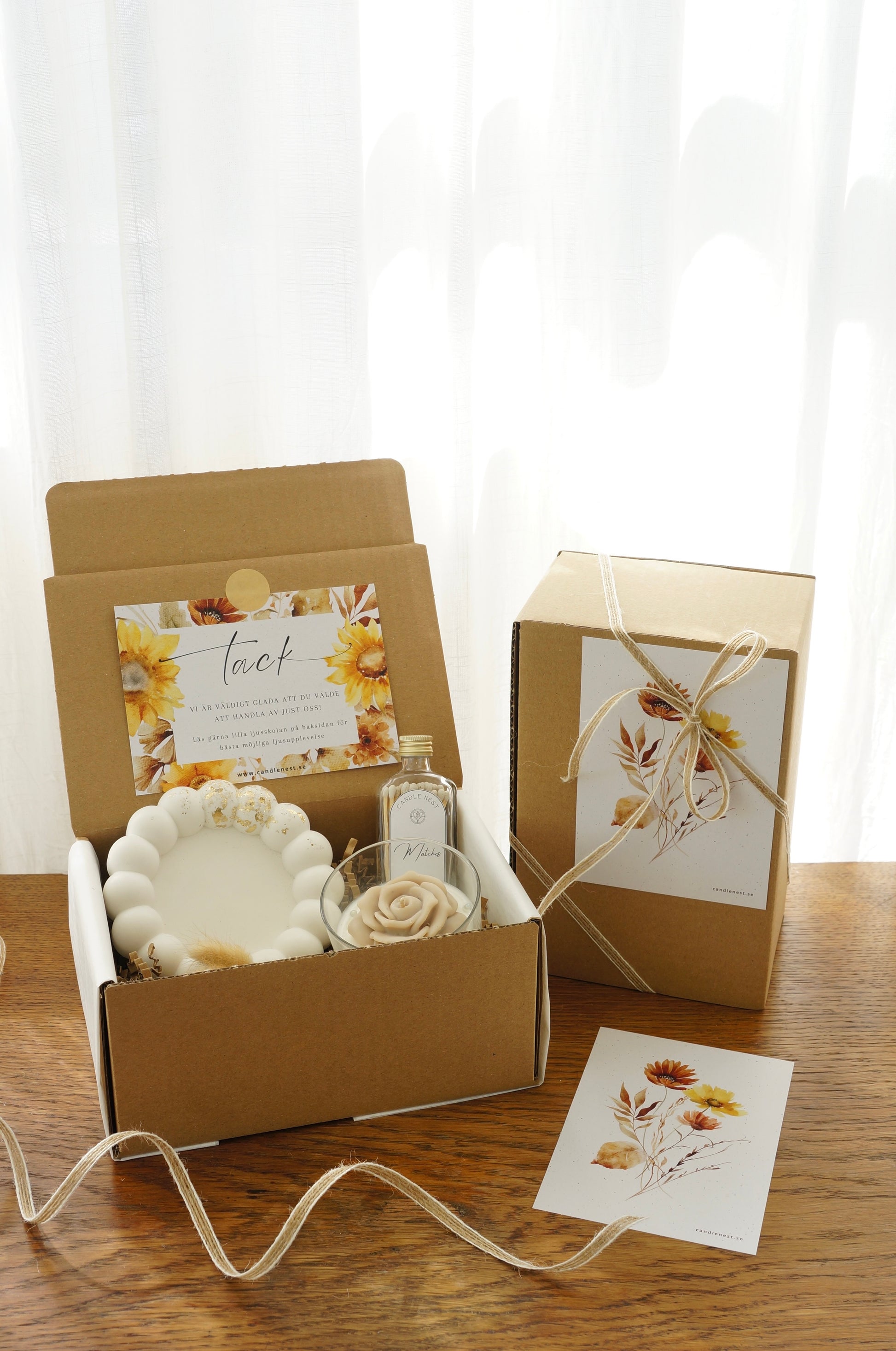 Lyxig Presentbox med ljusfat, Booms & Bubbles doftljus (Casablanca cocktail), flaska med tändstickor. Bild snett ovanifrån med en inslagen presentbox vid sidan