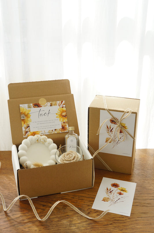 Lyxig Presentbox med ljusfat, Booms & Bubbles doftljus (Casablanca cocktail), flaska med tändstickor. Bild snett ovanifrån med en inslagen presentbox vid sidan
