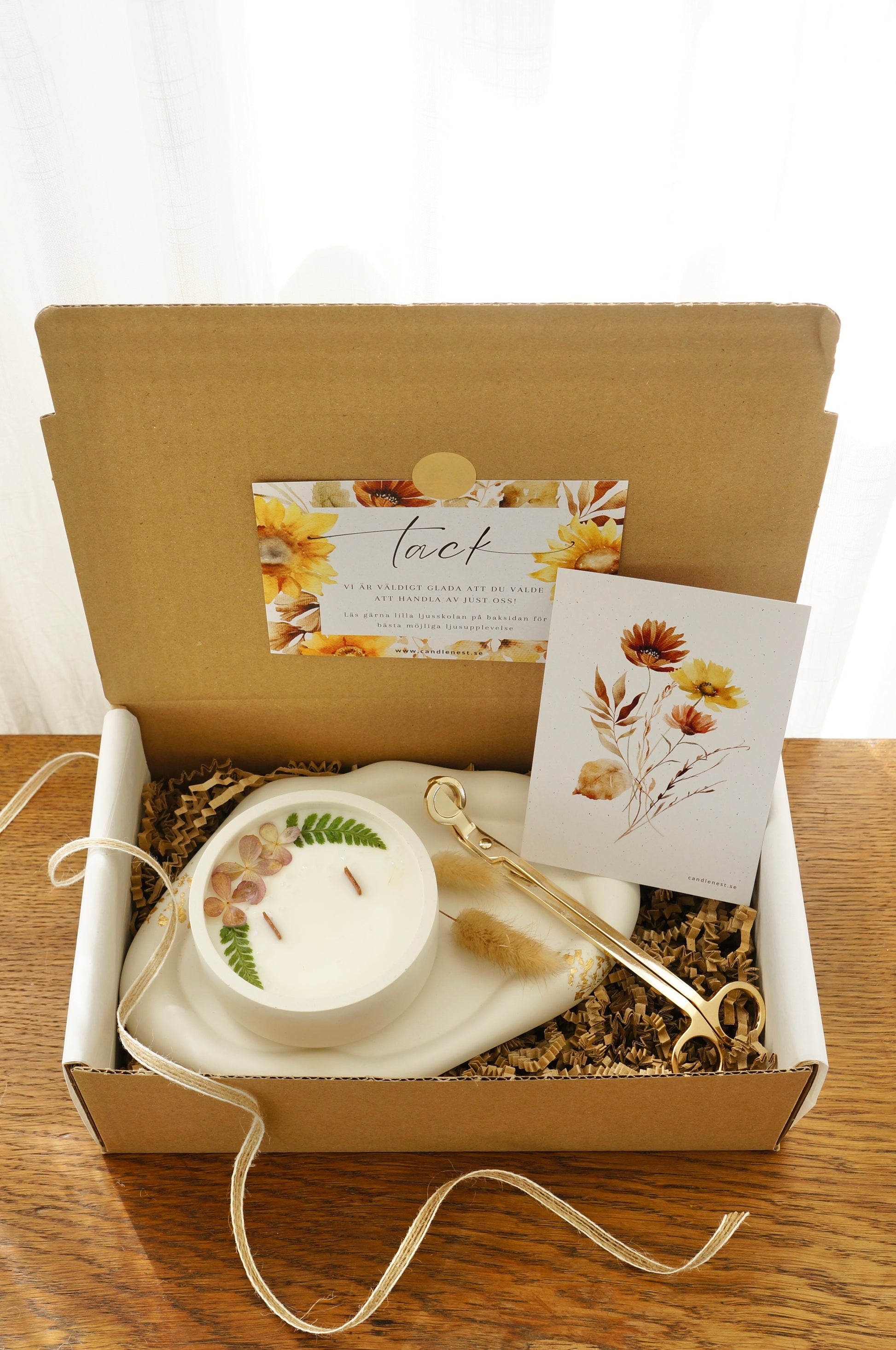 Presentbox med Botaniskt doftljus (spring morning walks), ljusfat med gyllen dekor och en vektång. Produkterna ligger i en låda av naturfärgd papp.