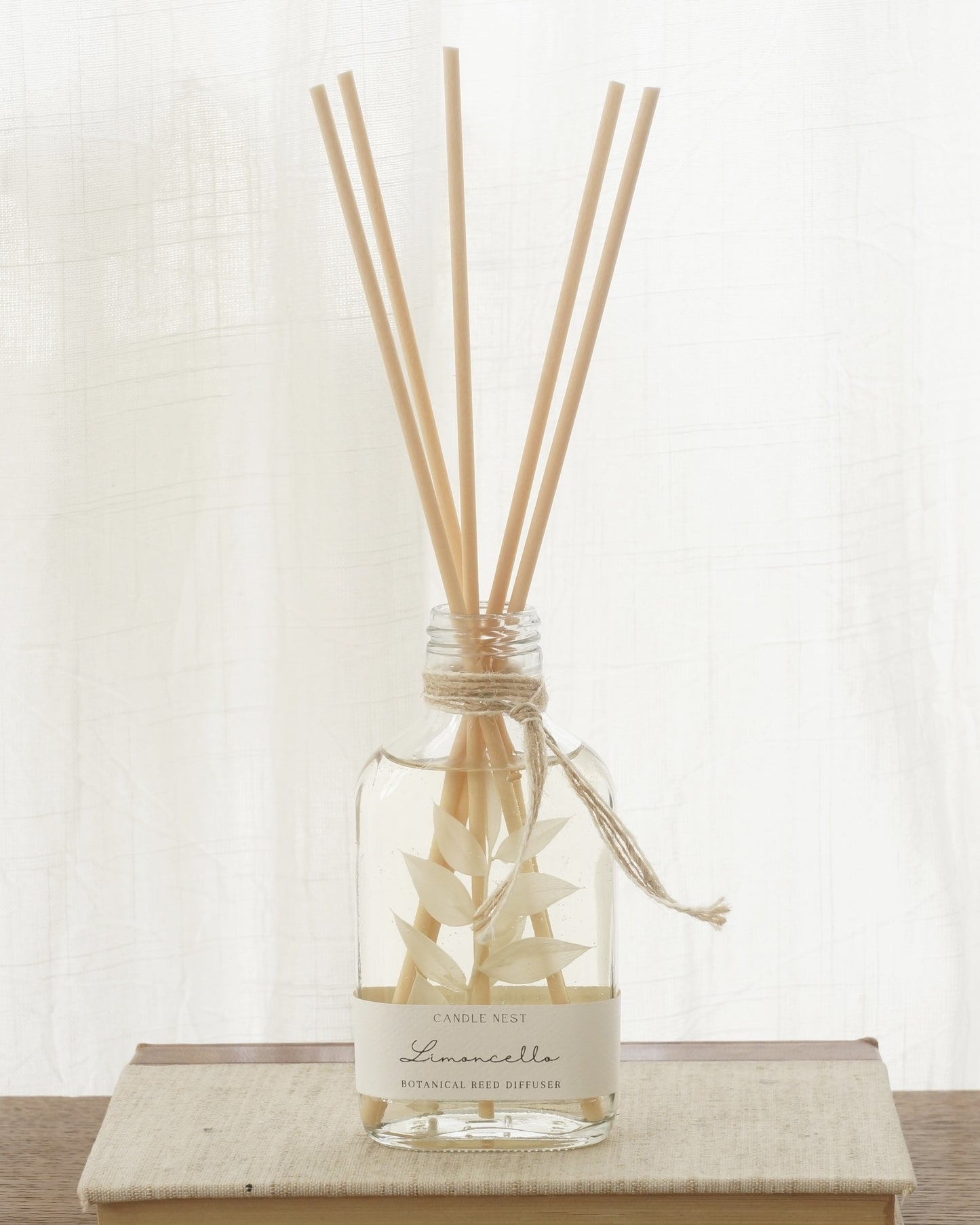 En genomskinlig flaska med doftpinnar står på ett bord. I flaskan syns en gren med vita blad. Flaskan har en skruvkork av guld.