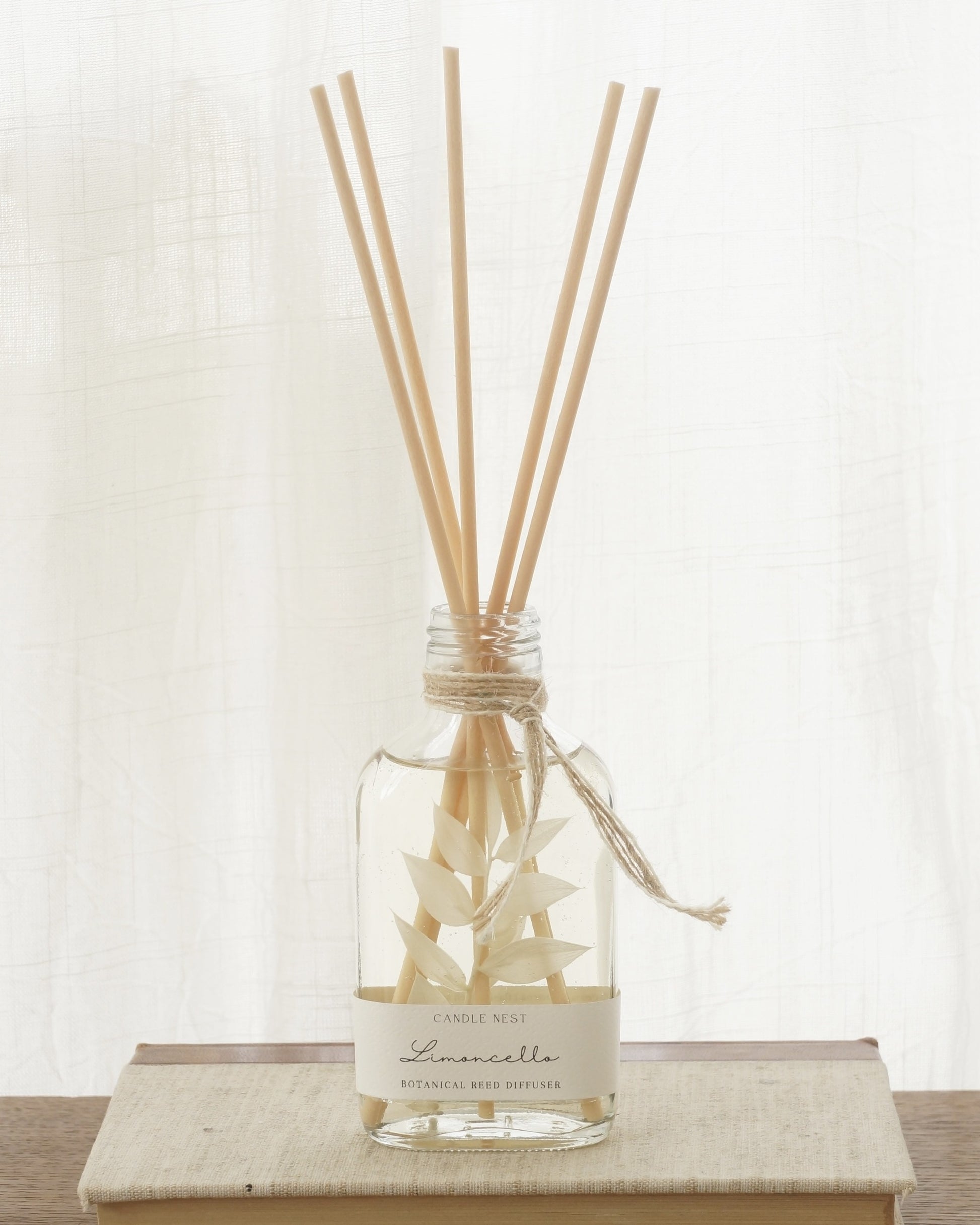 En genomskinlig flaska med doftpinnar står på ett bord. I flaskan syns en gren med vita blad. Flaskan har en skruvkork av guld.