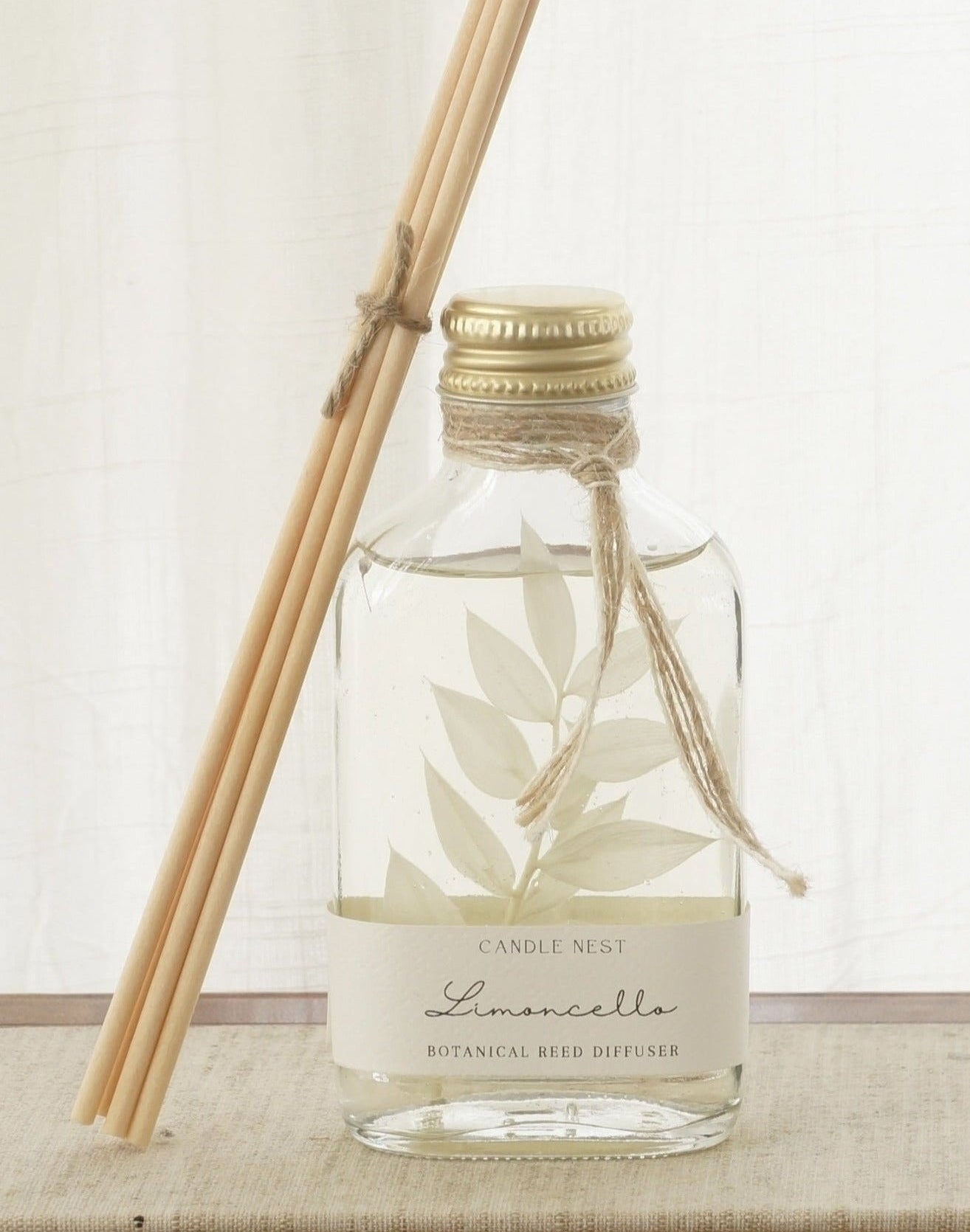 En genomskinlig flaska med doftpinnar står på ett bord. I flaskan syns en gren med vita blad. Flaskan har en skruvkork av guld och pinnarna (reeds)står lutade mot flaskan. 