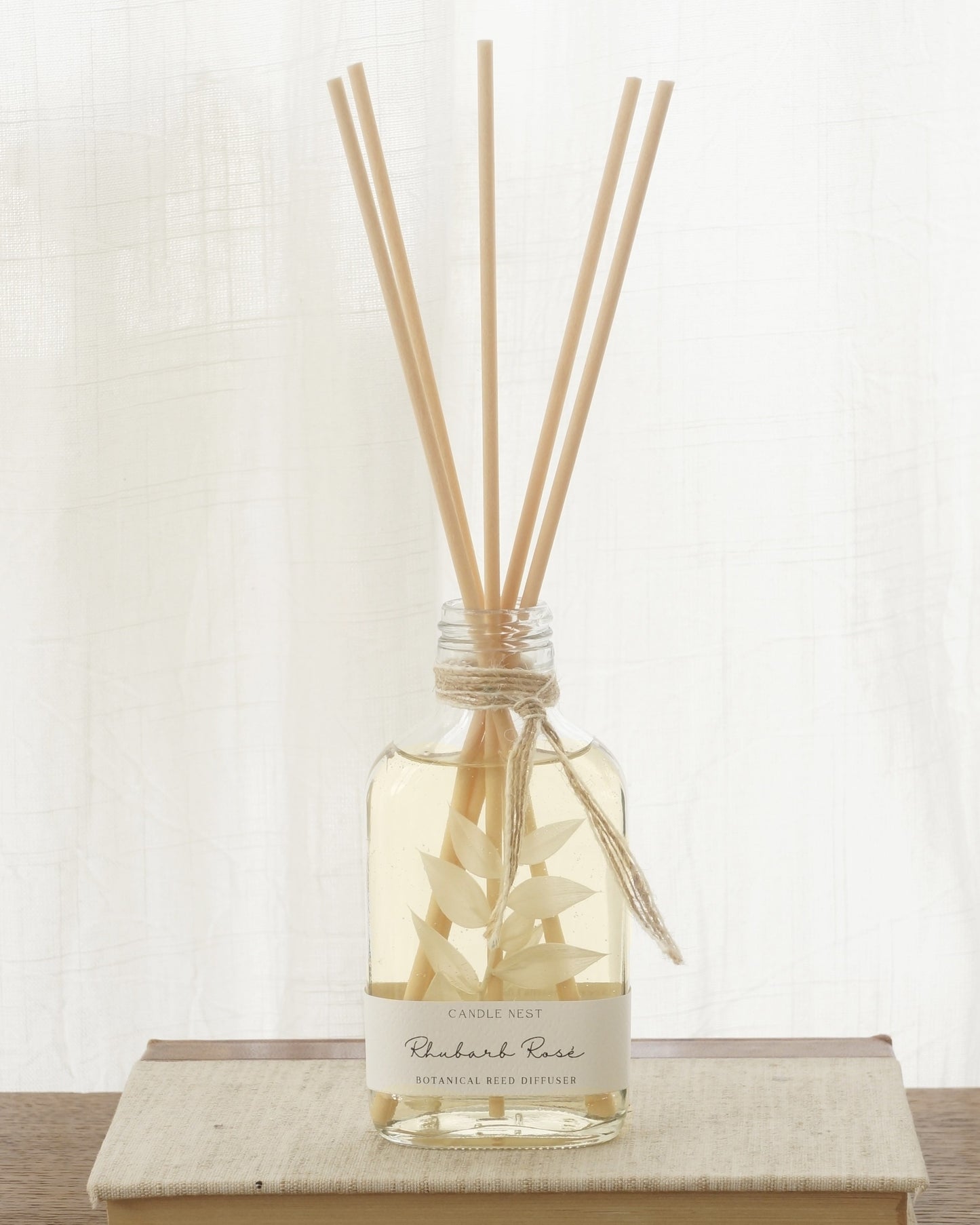 En genomskinlig flaska med doftpinnar står på ett bord. I flaskan syns en gren med vita blad. Flaskan har en skruvkork av guld.