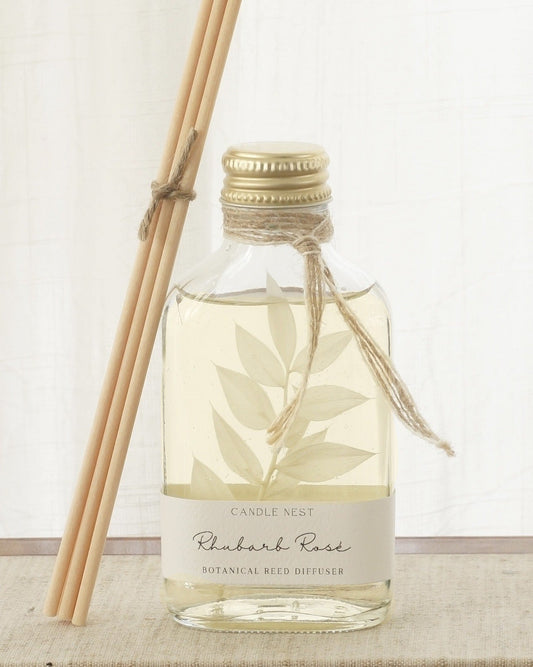 En genomskinlig flaska med doftpinnar står på ett bord. I flaskan syns en gren med vita blad. Flaskan har en skruvkork av guld och pinnarna (reeds)står lutade mot flaskan. 