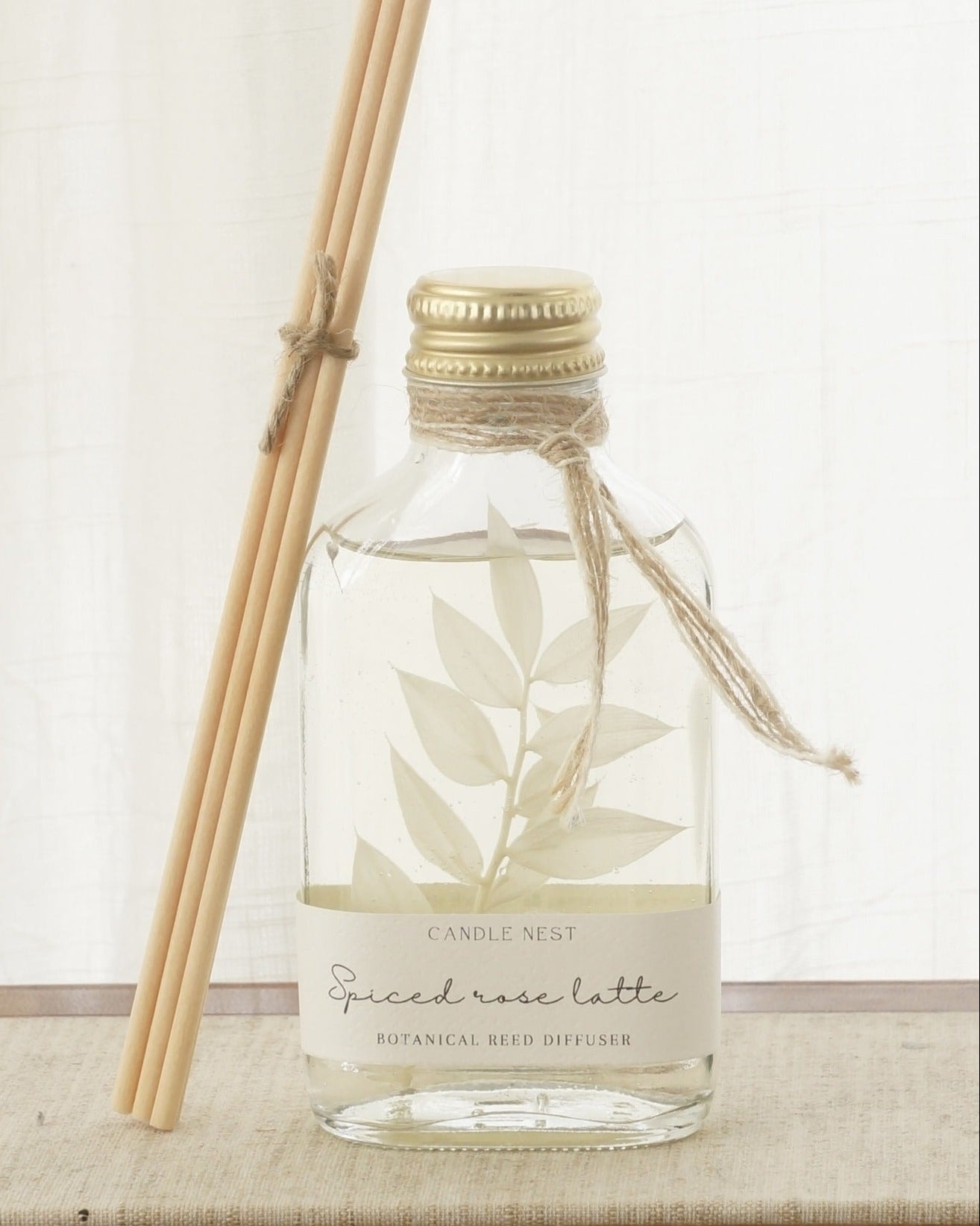 En genomskinlig flaska med doftpinnar står på ett bord. I flaskan syns en gren med vita blad. Flaskan har en skruvkork av guld och pinnarna (reeds)står lutade mot flaskan. 