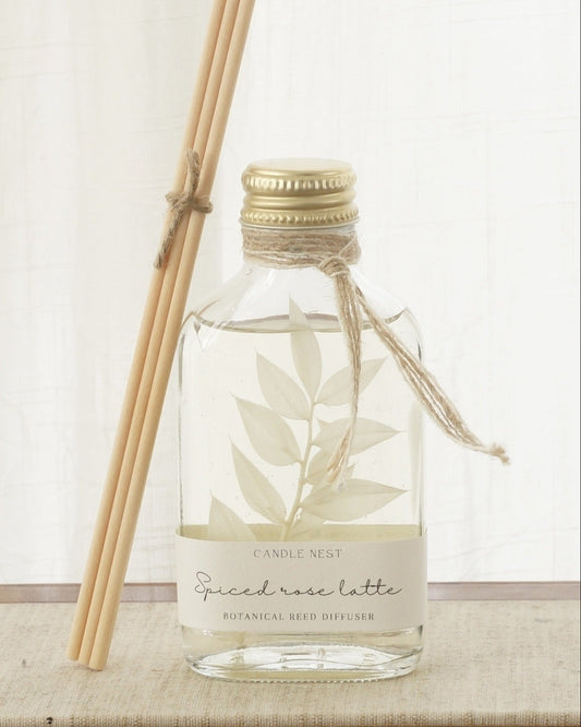 En genomskinlig flaska med doftpinnar står på ett bord. I flaskan syns en gren med vita blad. Flaskan har en skruvkork av guld och pinnarna (reeds)står lutade mot flaskan. 