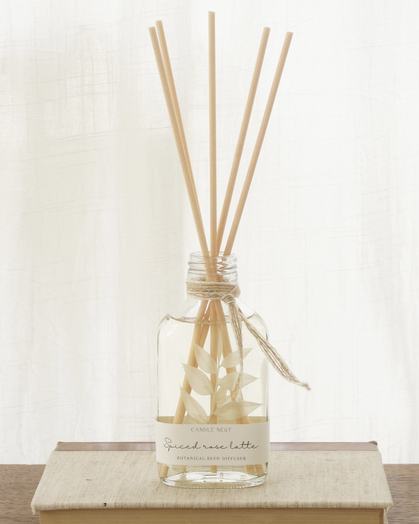 En genomskinlig flaska med doftpinnar står på ett bord. I flaskan syns en gren med vita blad. Flaskan har en skruvkork av guld.