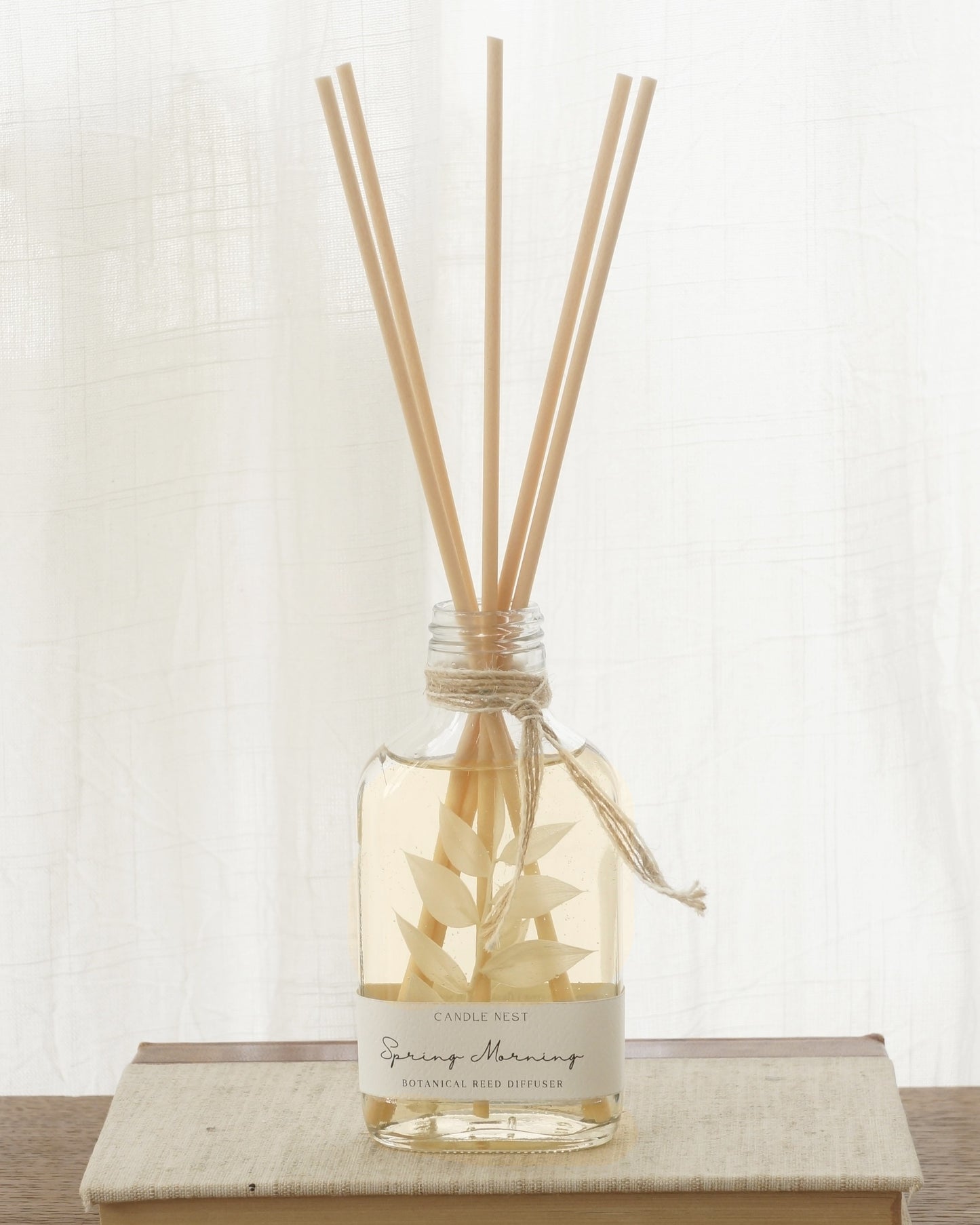 En genomskinlig flaska med doftpinnar står på ett bord. I flaskan syns en gren med vita blad. Flaskan har en skruvkork av guld.