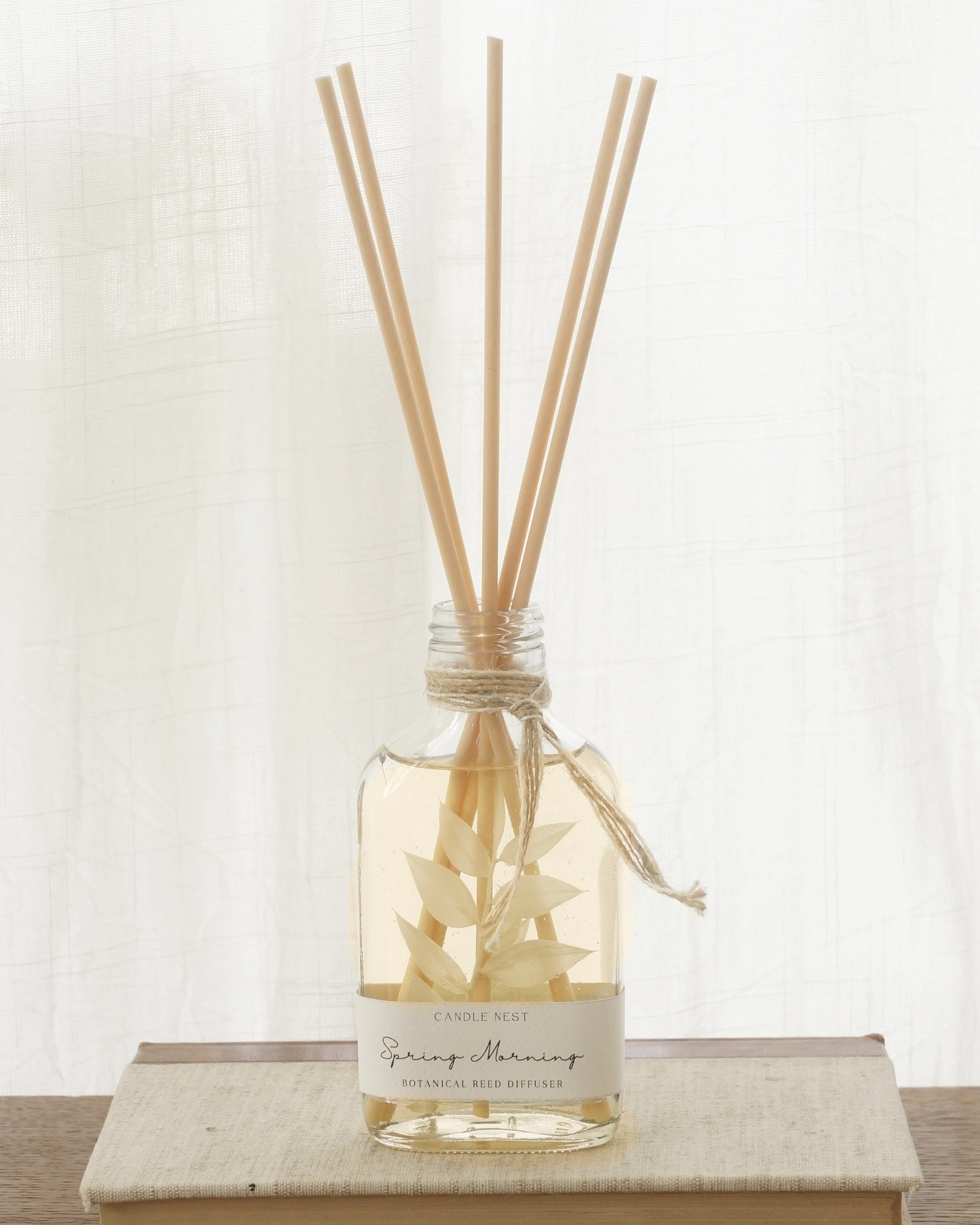 En genomskinlig flaska med doftpinnar står på ett bord. I flaskan syns en gren med vita blad. Flaskan har en skruvkork av guld.