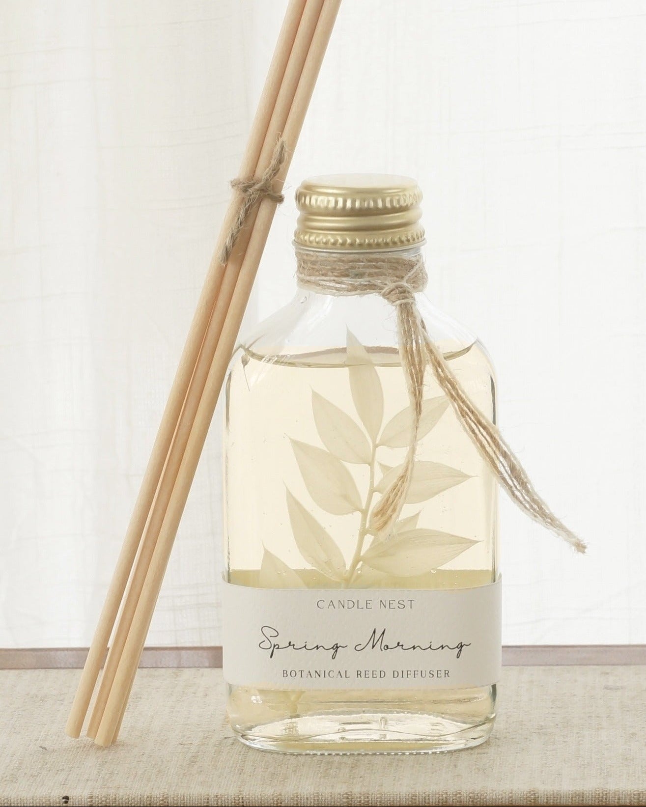 En genomskinlig flaska med doftpinnar står på ett bord. I flaskan syns en gren med vita blad. Flaskan har en skruvkork av guld och pinnarna (reeds)står lutade mot flaskan. 