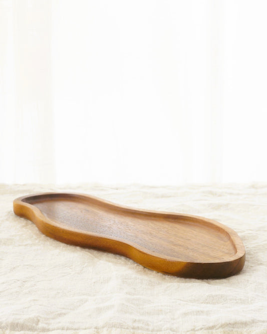 Avlångt träfat i akacia med mjukt rundade kanter, liggande på en ljus linneduk. Bild tagen från sidan