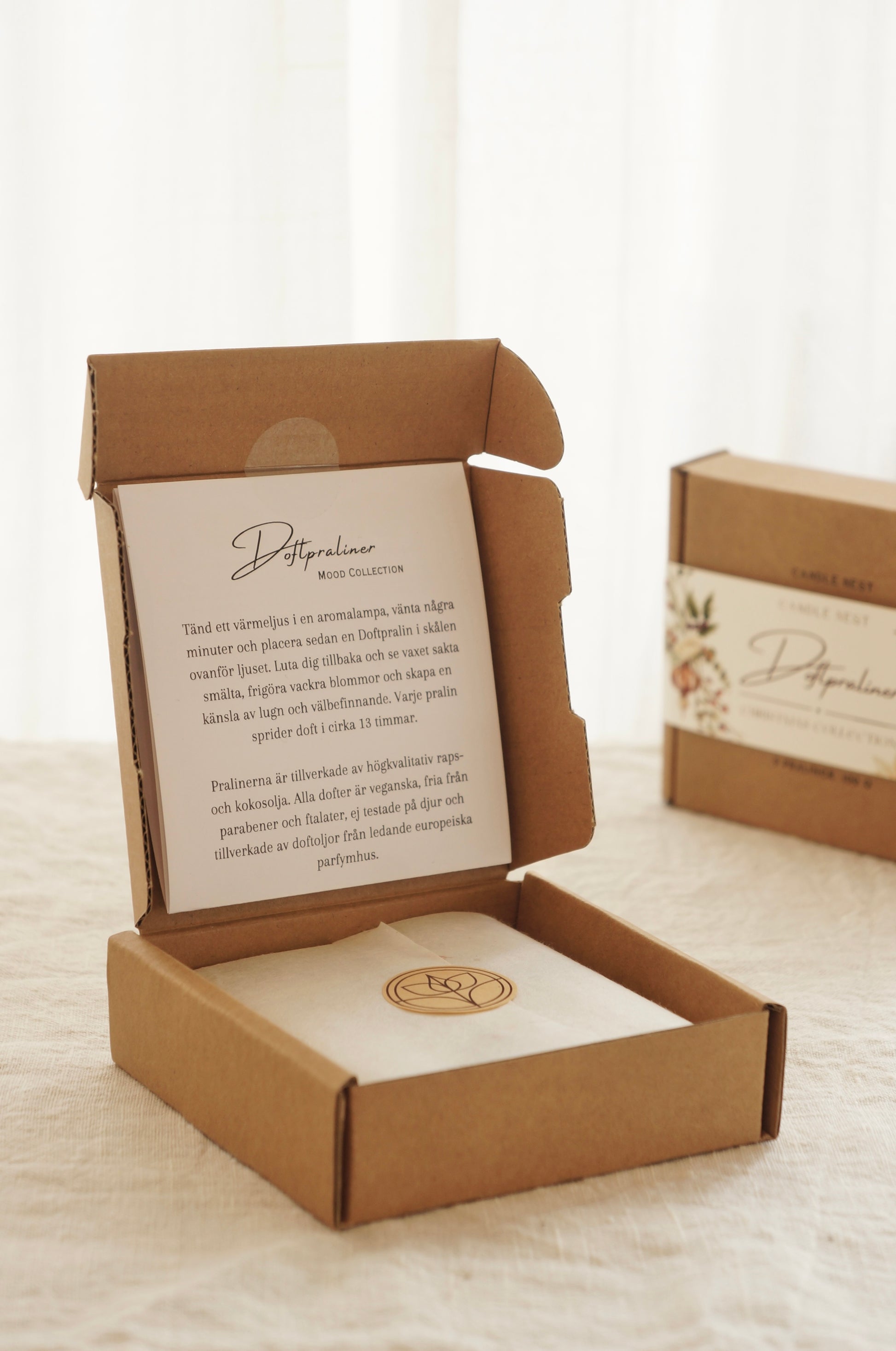En ask i naturfärgad papp innehållandes 9 st doftvax (doftpraliner) står på ett bord. Asken är öppen men pralinerna syns inte då silkespappret är slutet. 