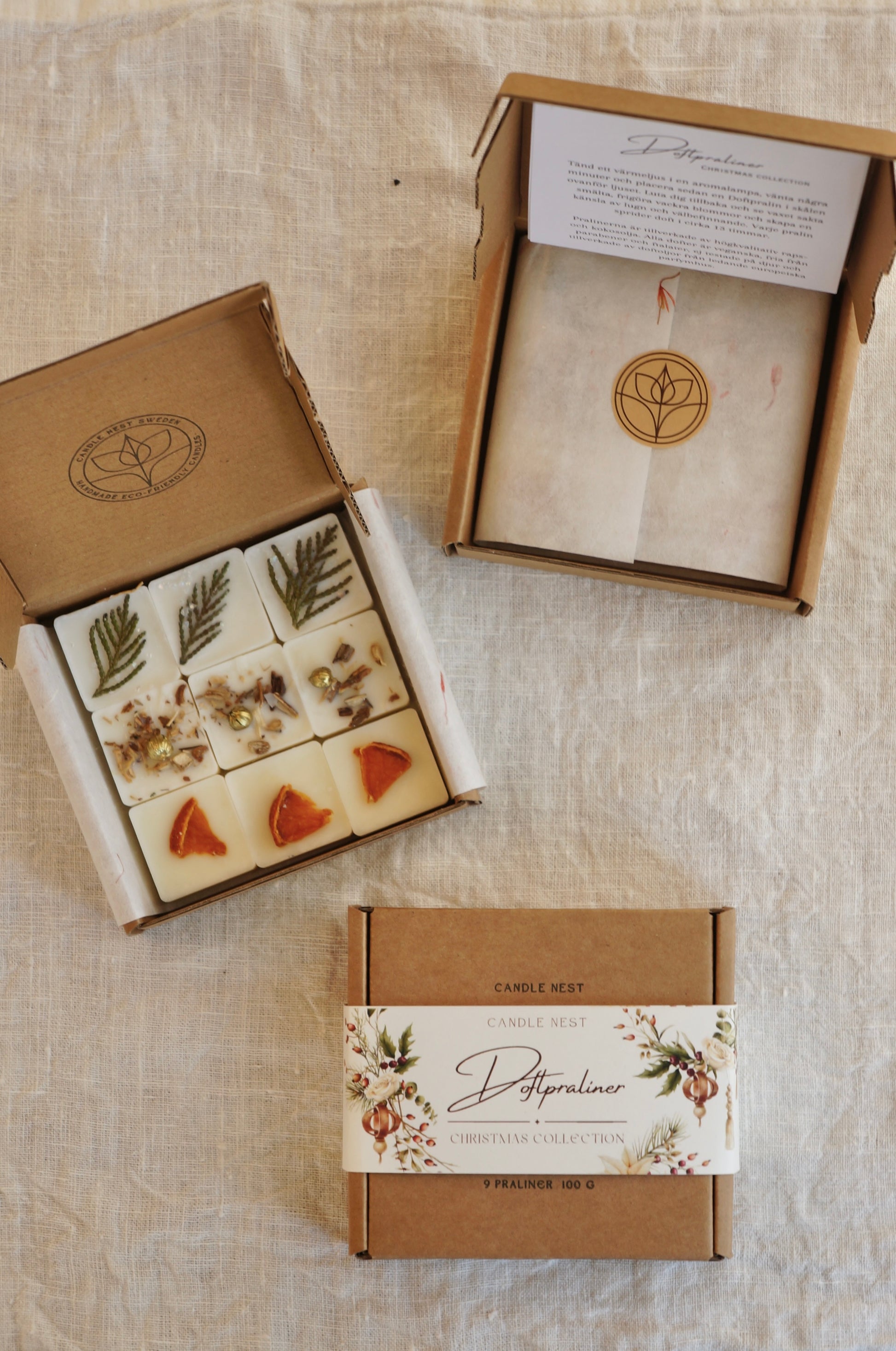 Tre askar i naturfärgad papp innehållandes 9 st doftvax (doftpraliner) ligger på ett bord. Doftpralinerna av raps och kokosvax är dekorerade med torkade bitar av clementin och små bitar av thuja. En ask är öppen och blottar pralinerna, en är stängd och man ser det fina trycket på utsidan av asken. En är öppen, men man ser endast silkespappret. 