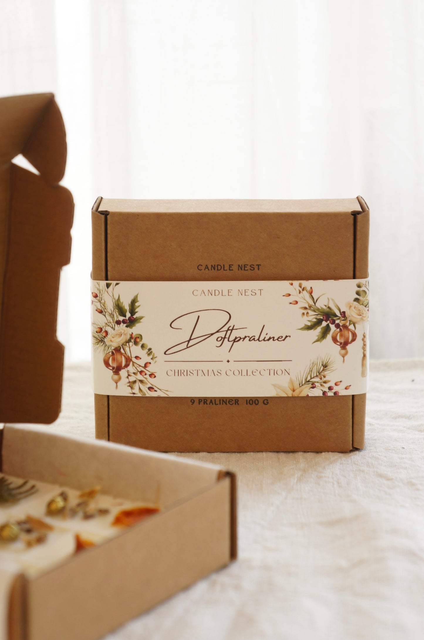 En ask i naturfärgad papp innehållandes 9 st doftvax (doftpraliner) står på ett bord när framsidan dan av asken syns. I förgrunden syns Doftpralinerna av raps och kokosvax är dekorerade med torkade bitar av clementin och små bitar av thuja. 
