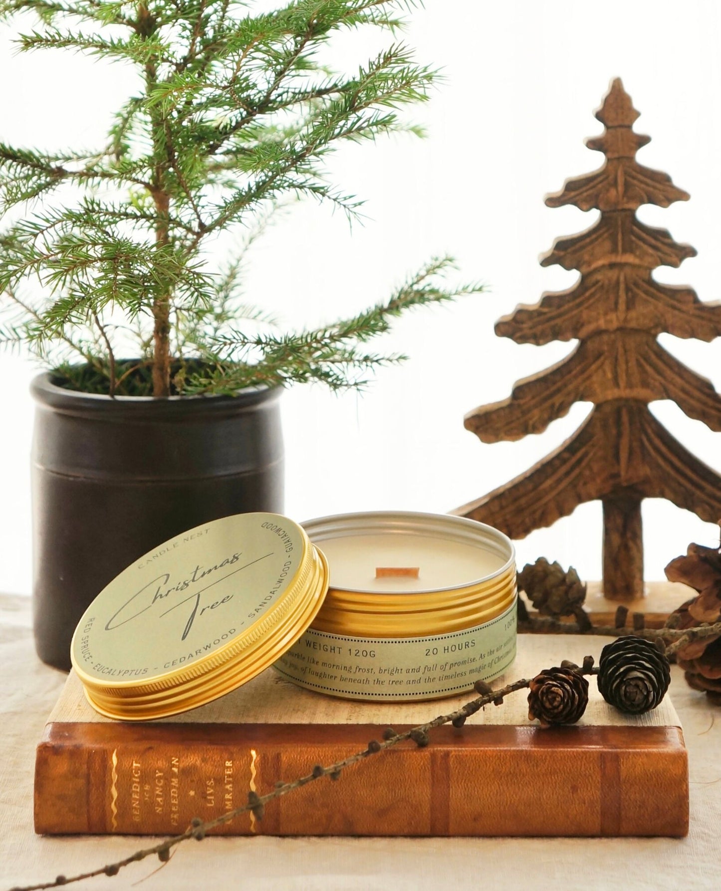 Ett doftljus i guldig aluminiumburk står på en äldre bok med rygg av läder. Mot burken står ljusets lock lutat. Mitt i ljuset syns en veke av trä. Bakom ljuset syns en urna med en Grankvist i samt en dekoration i trä som liknar en julgran. Runt och framför ljuset syns en kvist av lärkträd och Dess kottar  