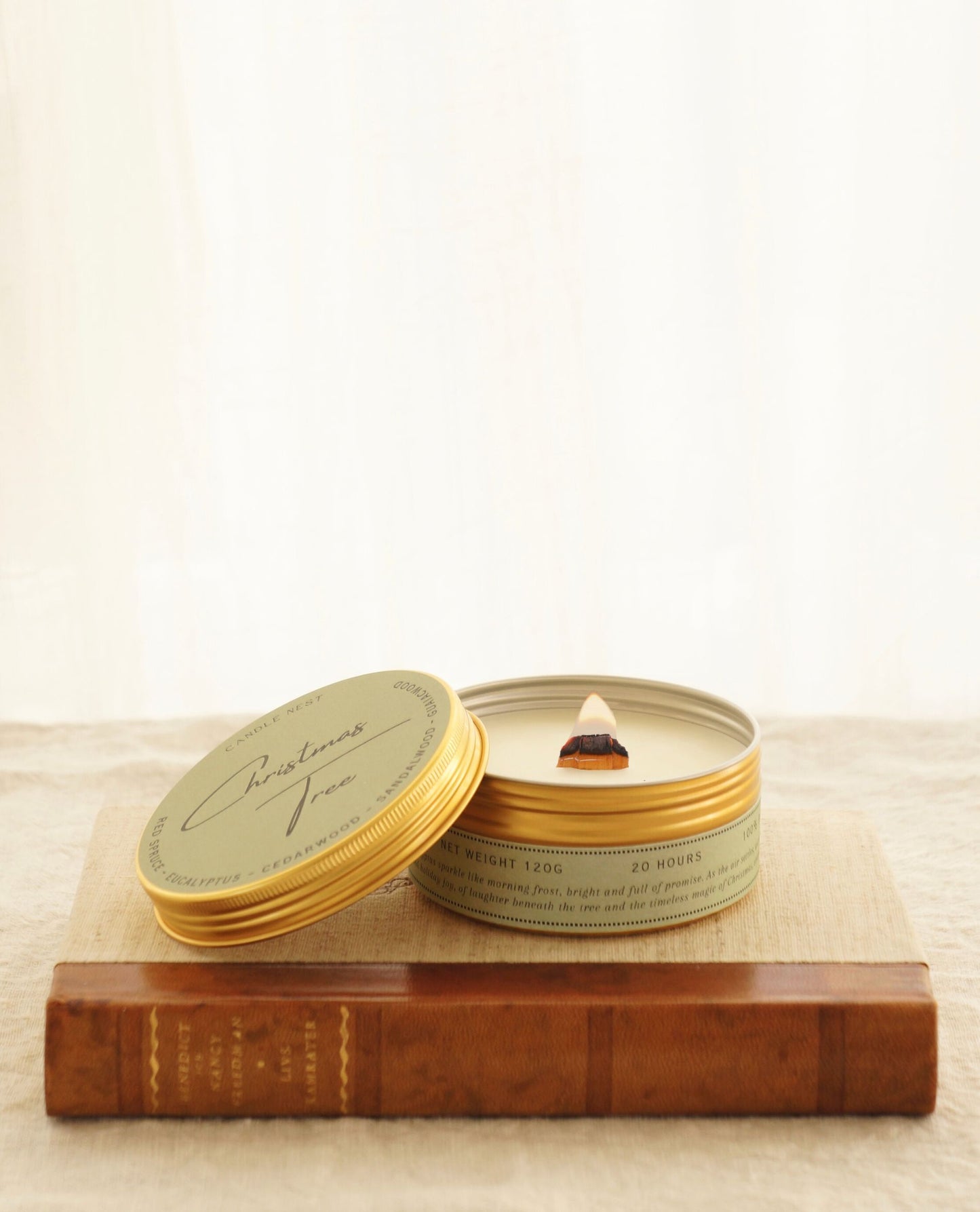 Ett doftljus i en guldig aluminiumburk står på en äldre bok med rygg av läder. Ljusets lock står lutat mot burken och på detta står det ” Christmas tree” med en snirklig vacker text. Mitt i ljuset syns en veke av trä. 