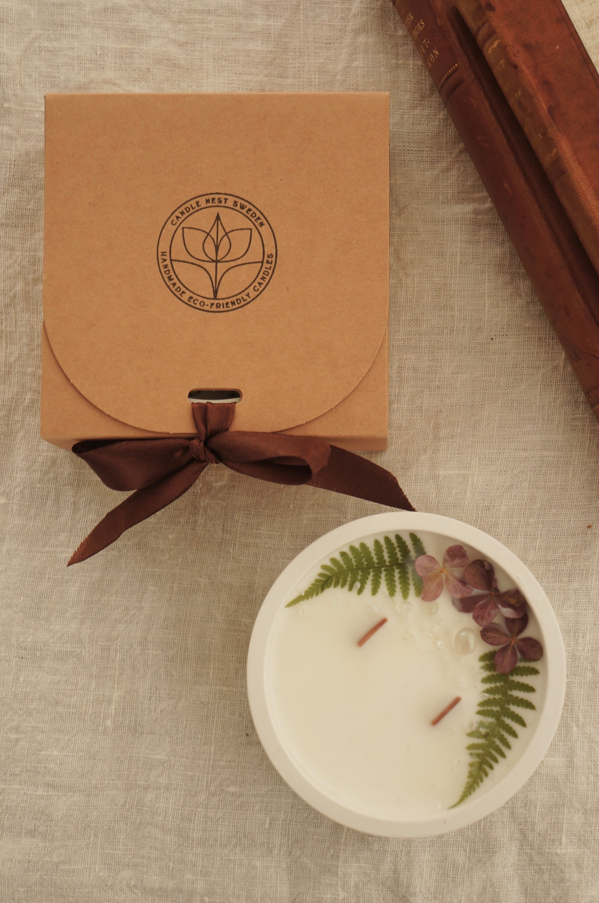 Ett runt doftljus i en vit skål av gips står på ett bord. Ljuset har två vekar av trä och ovansidan av ljuset är dekorerat med tre blommor av torkad hortensia, två små blad av ormbunke samt några glaspärlor. Bredvid ljuset står en naturfärgad ask med Candle Nests runda  logga på. Asken knyts ihop med ett brunt sidenband. Objekten är placerade på en beige duk av linnetyg och bilden är tagen uppifrån. 