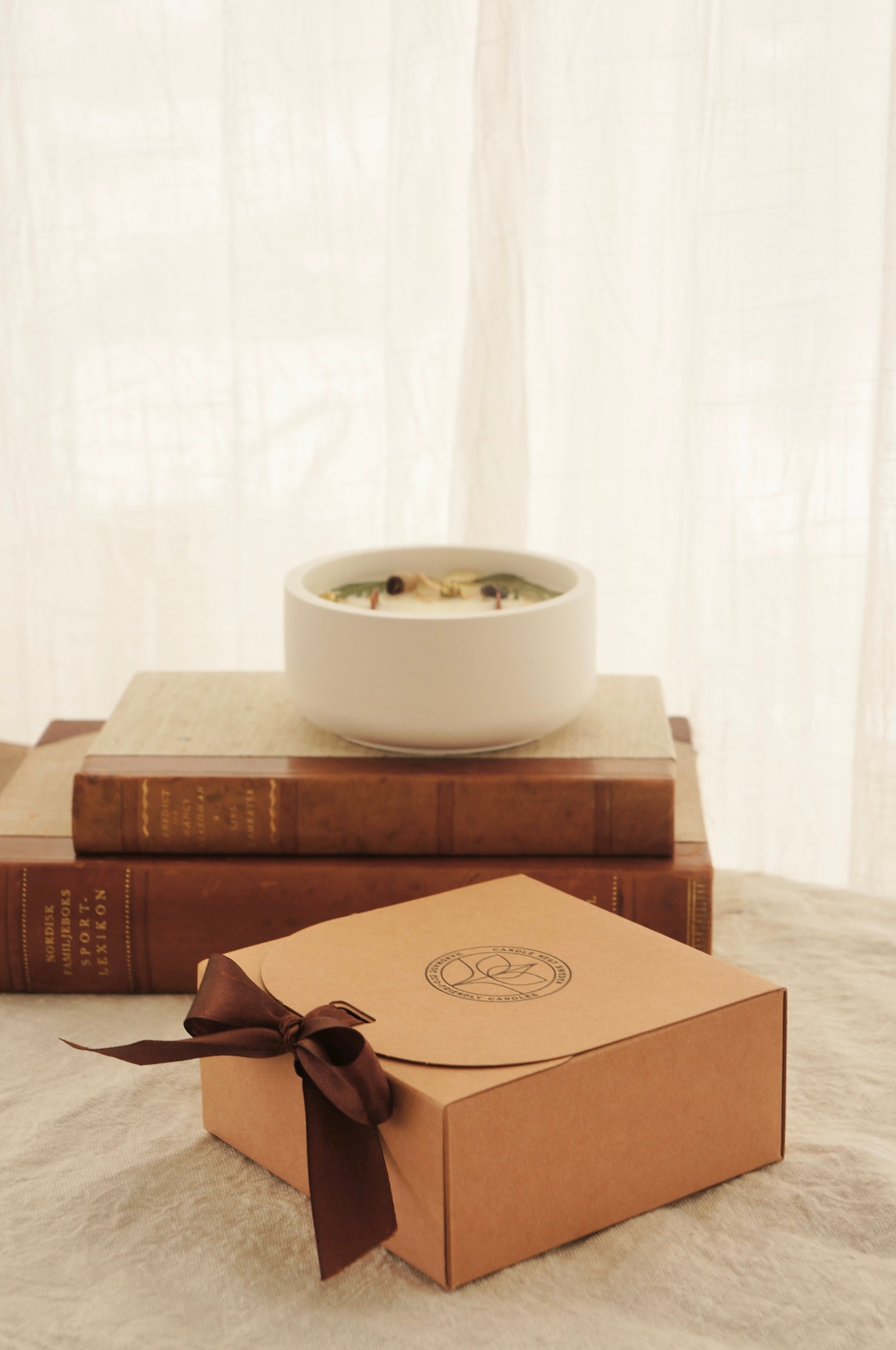 Bild i varma färger. En ask i naturfärgad papp står på ett bord. Ett brunt sidenband håller locket på plats. Bakom asken syns två äldre böcker med läderrygg. På böckerna står ett doftljus i en vit gipsskål. 