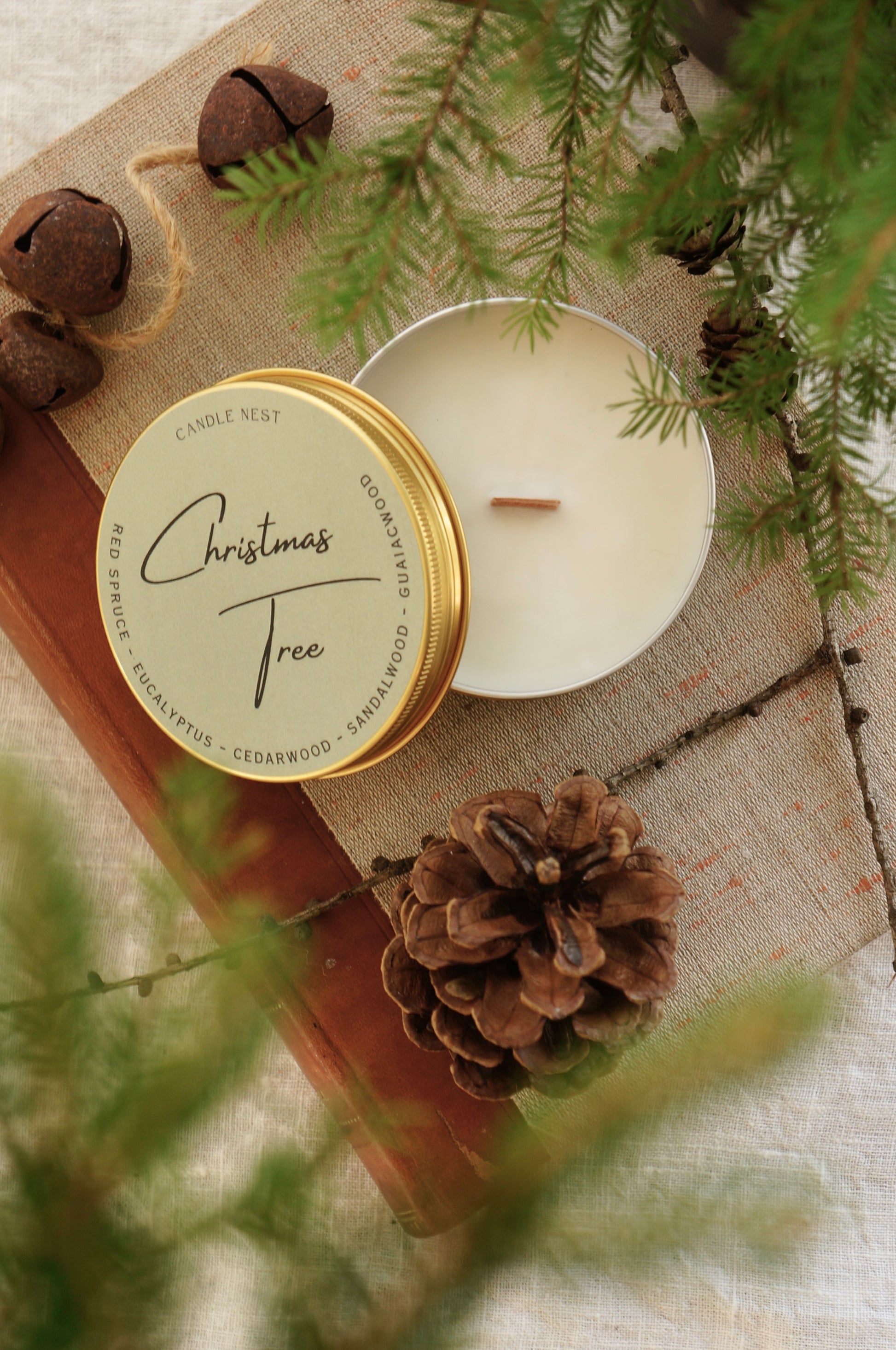 Ett doftljus i en guldig aluminiumburk står på en äldre bok med rygg av läder. Locket är lutat mot burken och på detta finns ” Christmas tree” skrivit med snirklig text. Ljuset har en veke av trä. Runt boken syns kottar och granris. 