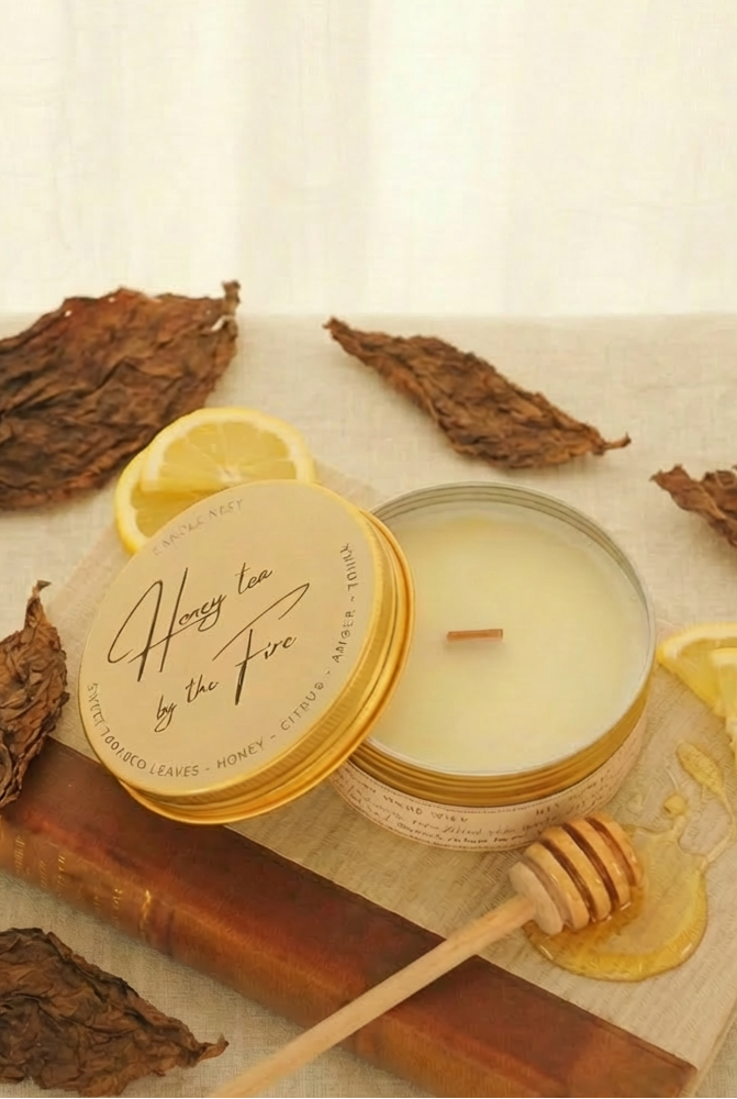 Ett doftljus i en rund guldfärgad burk av metall står på en bok. Boken är av linnetyg med en rygg i läder. Ljusets lock står lutad på ljuset på på det finns ljusets namn "Honey tea at the Fire" skrivet med snirklig text. Mitt i ljuset syns en veke av trä. Runt ljuset ligger torkade tobaksblad, och skivor av citron. På boken ligger en honungsstav och lite honung flyter ut på boken. Bilden är tagen snett uppifrån.