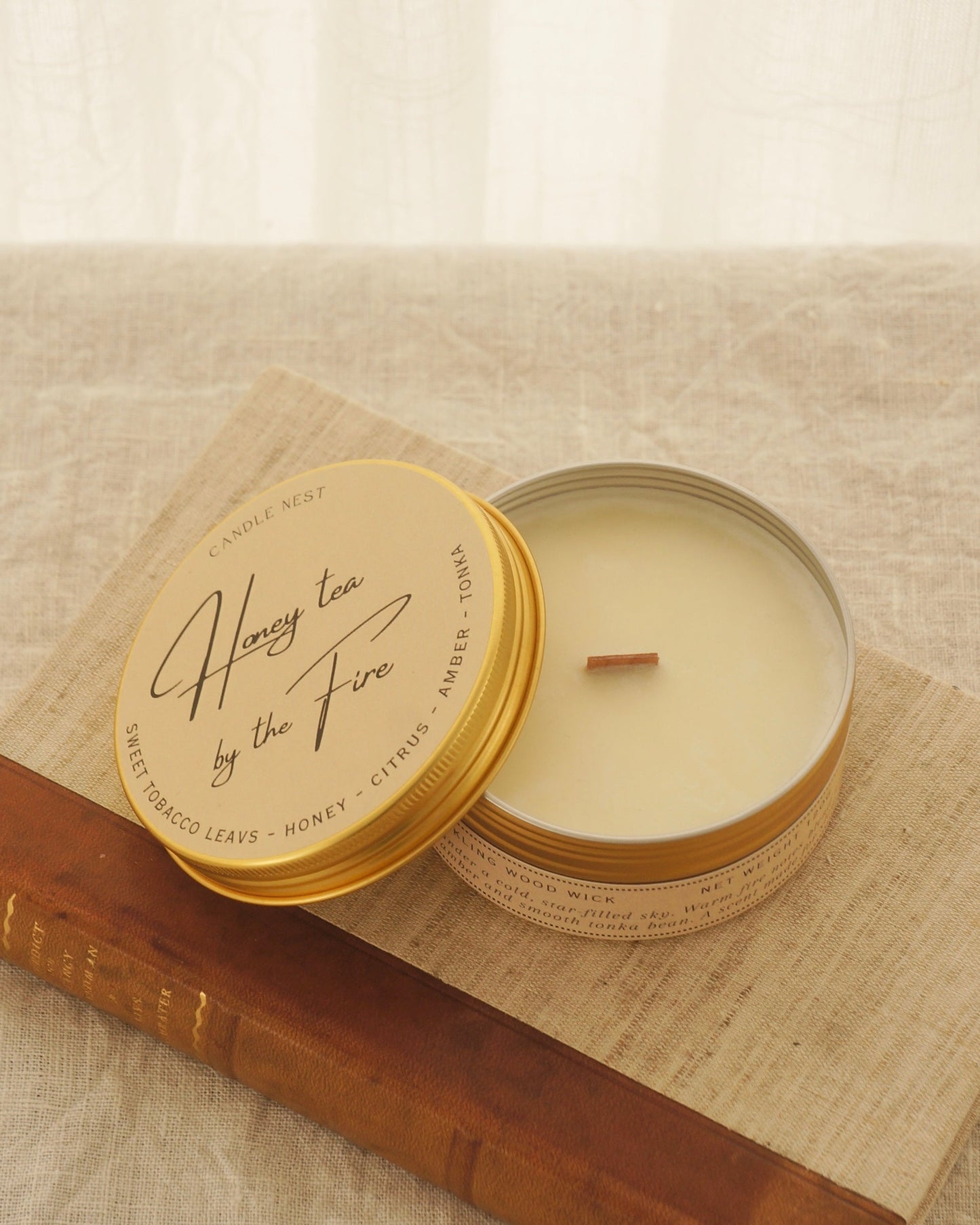 Ett doftljus i en rund guldfärgad burk av metall står på en bok. Boken är av linnetyg med en rygg i läder. Ljusets lock står lutad på ljuset på på det finns ljusets namn "Honey tea at the Fire" skrivet med snirklig text. Mitt i ljuset syns en veke av trä. Bilden är tagen snett uppifrån.