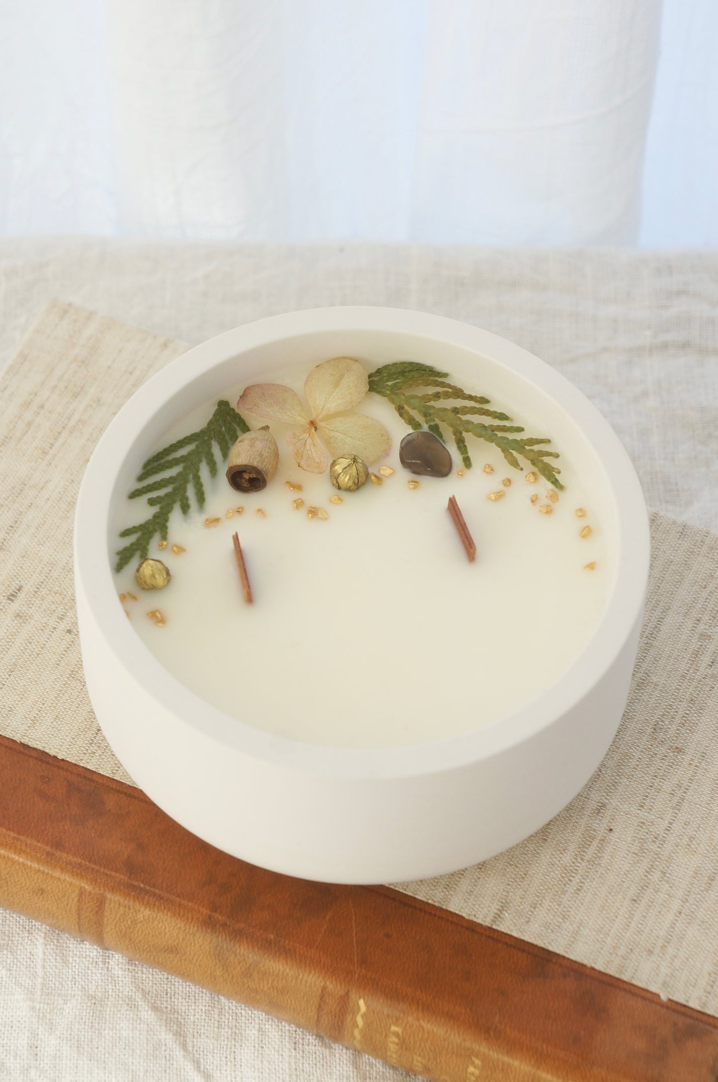 Ett doftljus i en rund vit skål av gips står på en bok. Boken är av linnetyg med en rygg i läder. Boken står på en beige duk av linnetyg. Mitt i ljuset syns två vekar av trä. Kanterna av ljusets yta är dekorerat med torkade växter. Inga växter finns nära vekarna. Bilden är tagen snett uppifrån.