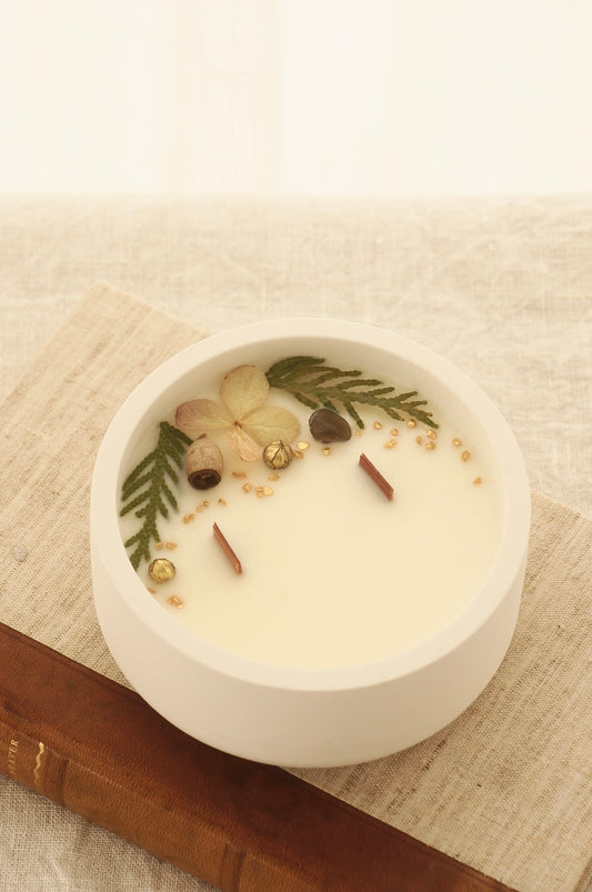 Ett doftljus i en rund vit skål av gips står på en bok. Boken är av linnetyg med en rygg i läder. Boken står på en beige duk av linnetyg. Mitt i ljuset syns två vekar av trä. Kanterna av ljusets yta är dekorerat med torkade växter. Inga växter finns nära vekarna. Bilden är tagen snett uppifrån.