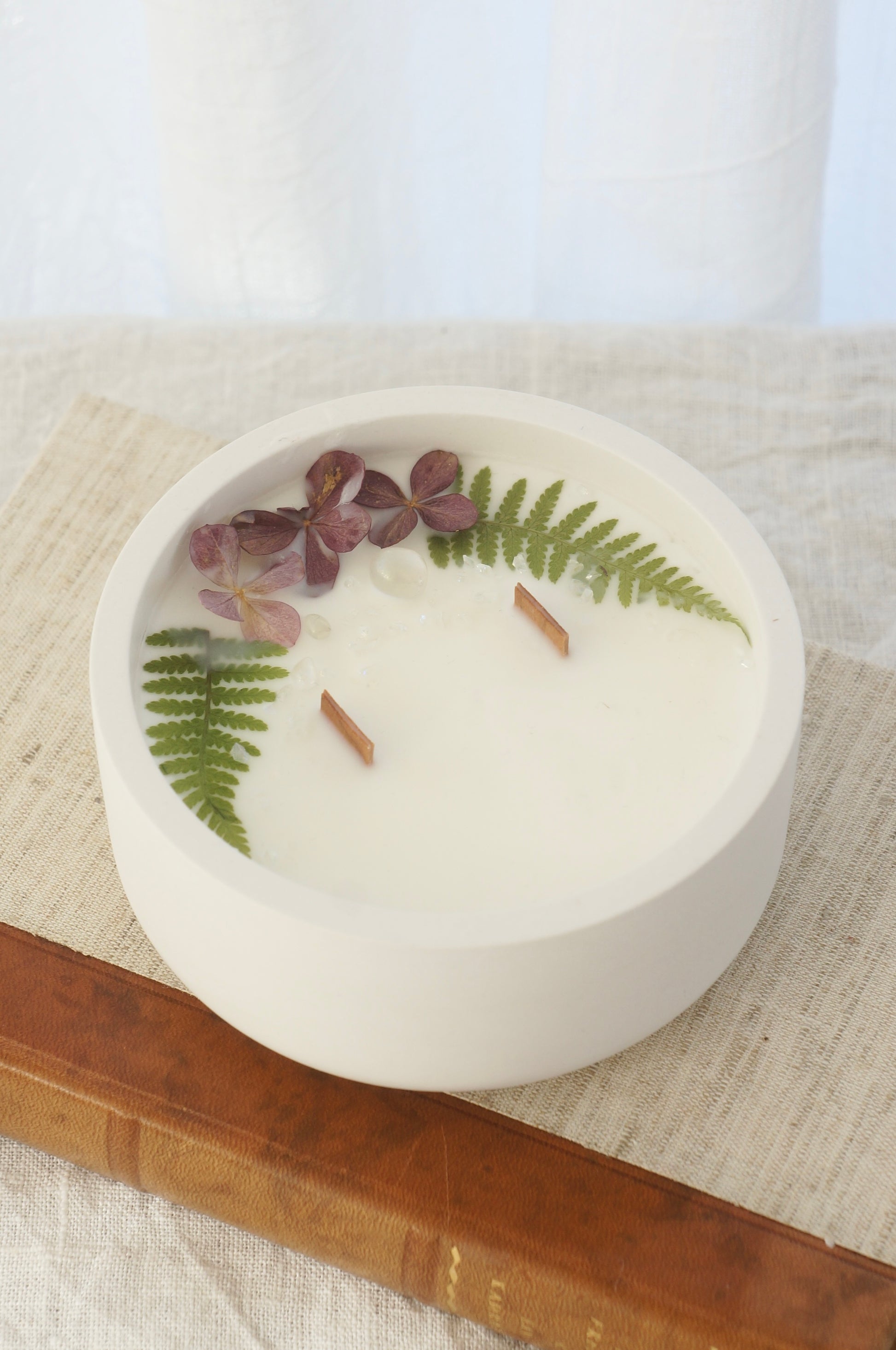 Ett doftljus i en rund vit skål av gips står på en bok. Boken är av linnetyg med en rygg i läder. Boken står på en beige duk av linnetyg. Mitt i ljuset syns två vekar av trä. Kanterna av ljusets yta är dekorerat med torkade växter. Inga växter finns nära vekarna. Bilden är tagen snett uppifrån.