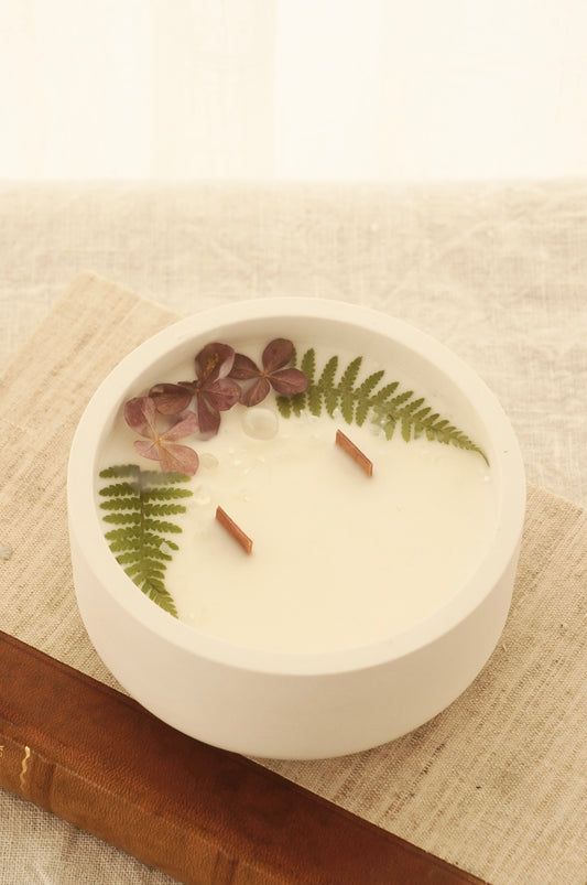 Ett doftljus i en rund vit skål av gips står på en bok. Boken är av linnetyg med en rygg i läder. Boken står på en beige duk av linnetyg. Mitt i ljuset syns två vekar av trä. Kanterna av ljusets yta är dekorerat med torkade växter. Inga växter finns nära vekarna. Bilden är tagen snett uppifrån.