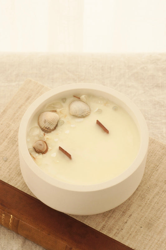 Ett doftljus i en rund vit skål av gips står på en bok. Boken är av linnetyg med en rygg i läder. Boken står på en beige duk av linnetyg. Mitt i ljuset syns två vekar av trä. Kanterna av ljusets yta är dekorerat med torkade växter. Inga växter finns nära vekarna. Bilden är tagen snett uppifrån.