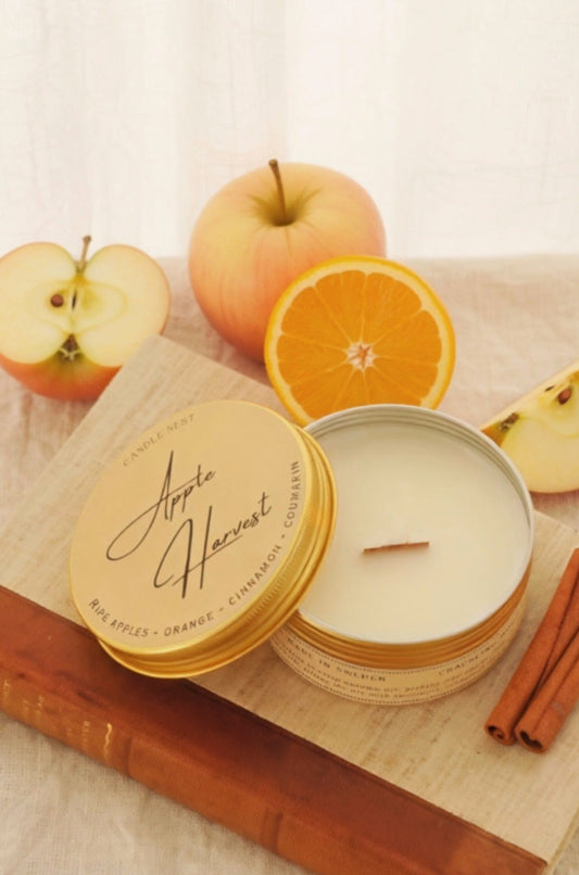 Ett doftljus i en rund guldfärgad burk av metall står på en bok. Boken har pärmar av av linnetyg med en rygg i läder. Ljusets lock står lutad på ljuset på på det finns ljusets namn skrivet med snirklig text. Mitt i ljuset syns en veke av trä. Boken ligger på en duk av linnetyg. Runt ljuset syns röda äpplen, en halv apelsin och två kanelstänger. Bilden är tagen snett uppifrån.